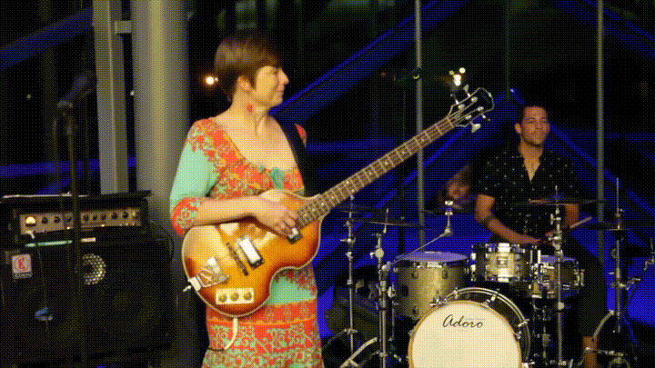Two musicians performing live, one playing electric guitar and the other on drums, in an indoor venue with large windows and colorful lighting.