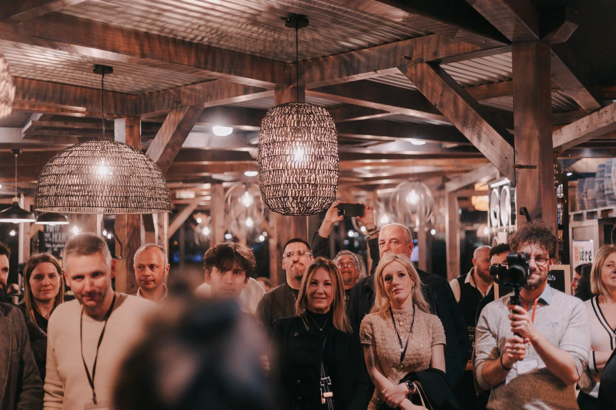 A crowded indoor event with wooden beams on the ceiling, pendant lights, and many people standing and watching something. One person is taking a photo with a camera, and another is holding a phone.