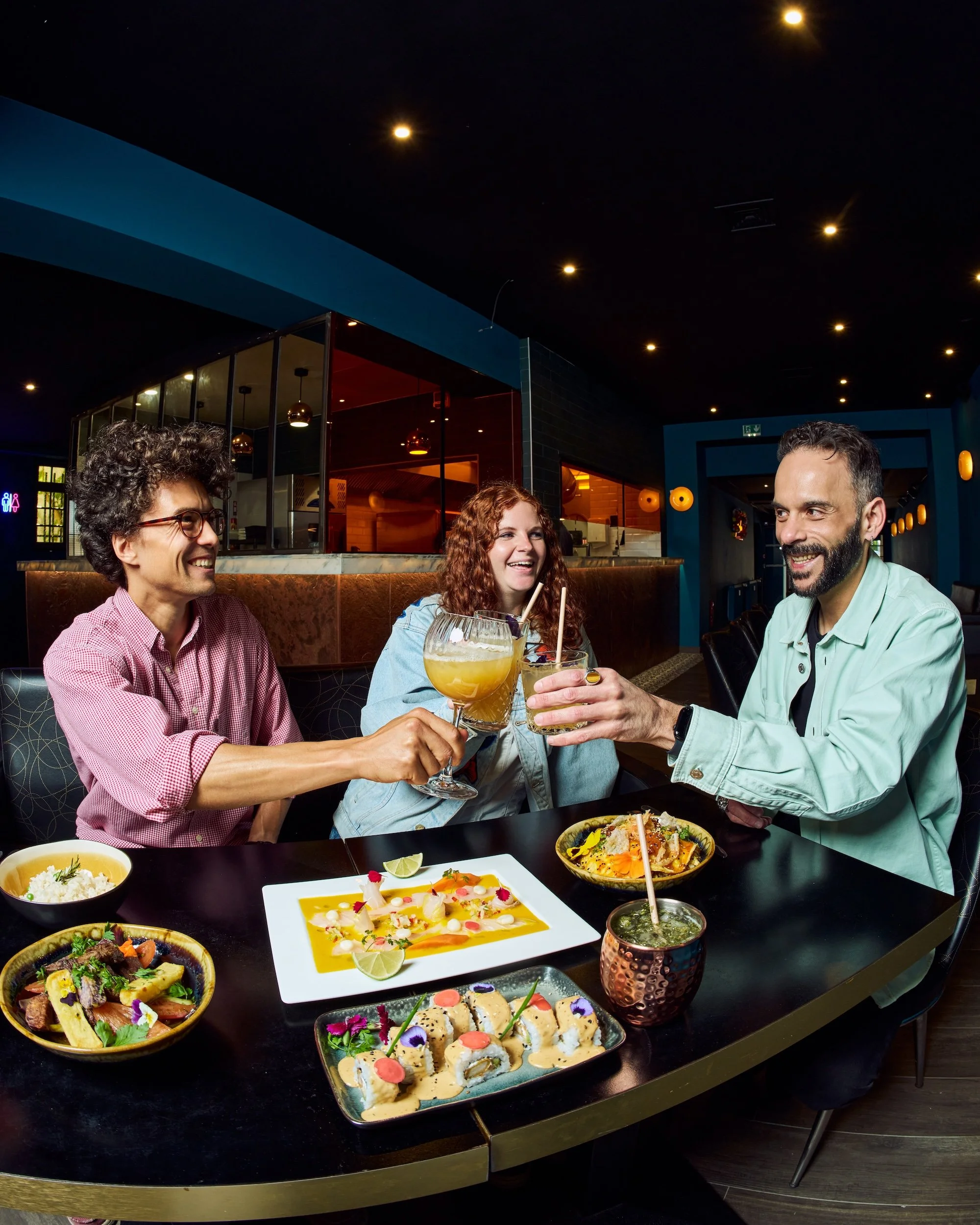 Three people smiling and toasting with cocktails at a restaurant table, surrounded by sushi dishes and other Asian foods.