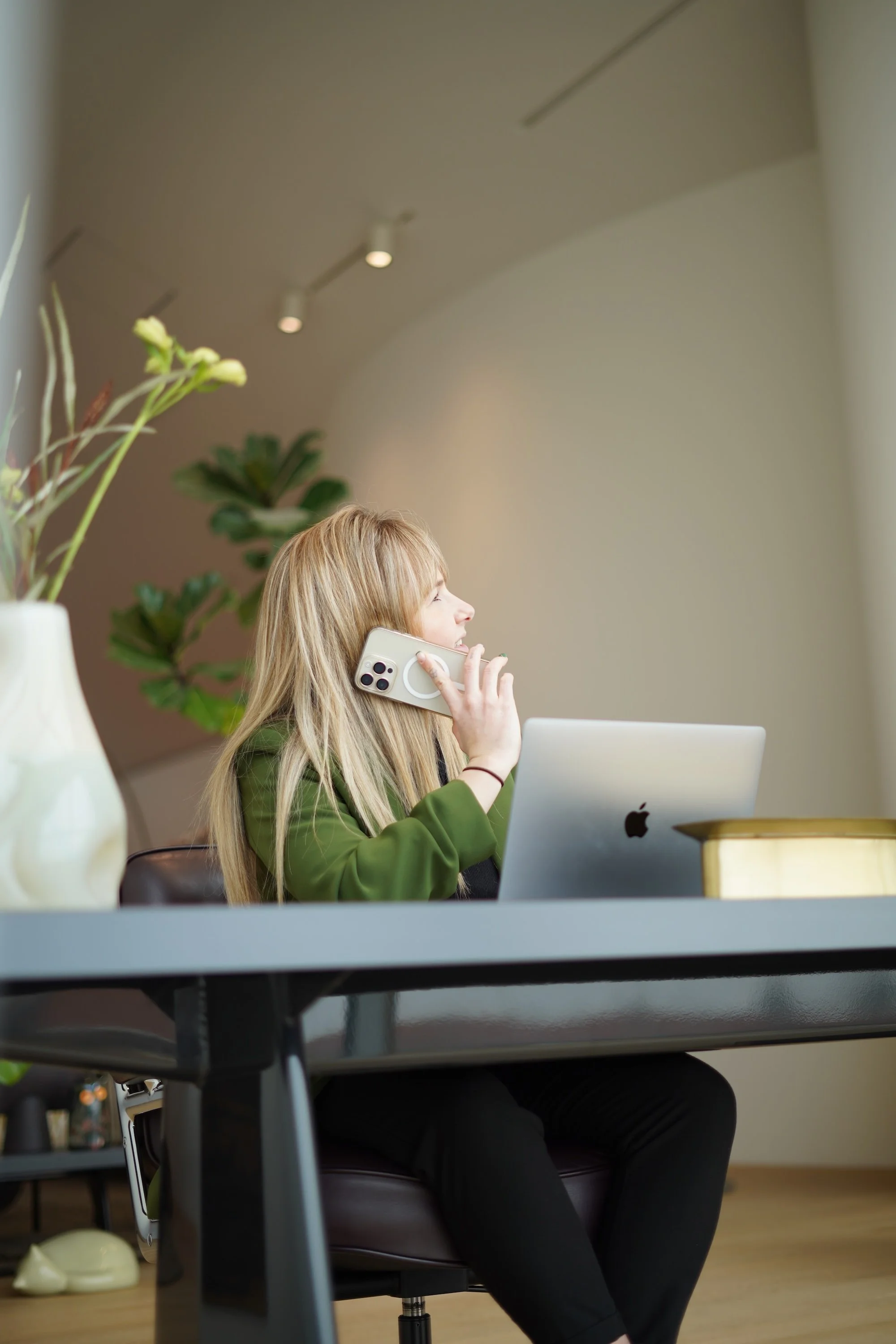 A woman with blonde hair, wearing a green jacket, sitting at a desk, talking on a smartphone, with a laptop in front of her and green plants in the background.