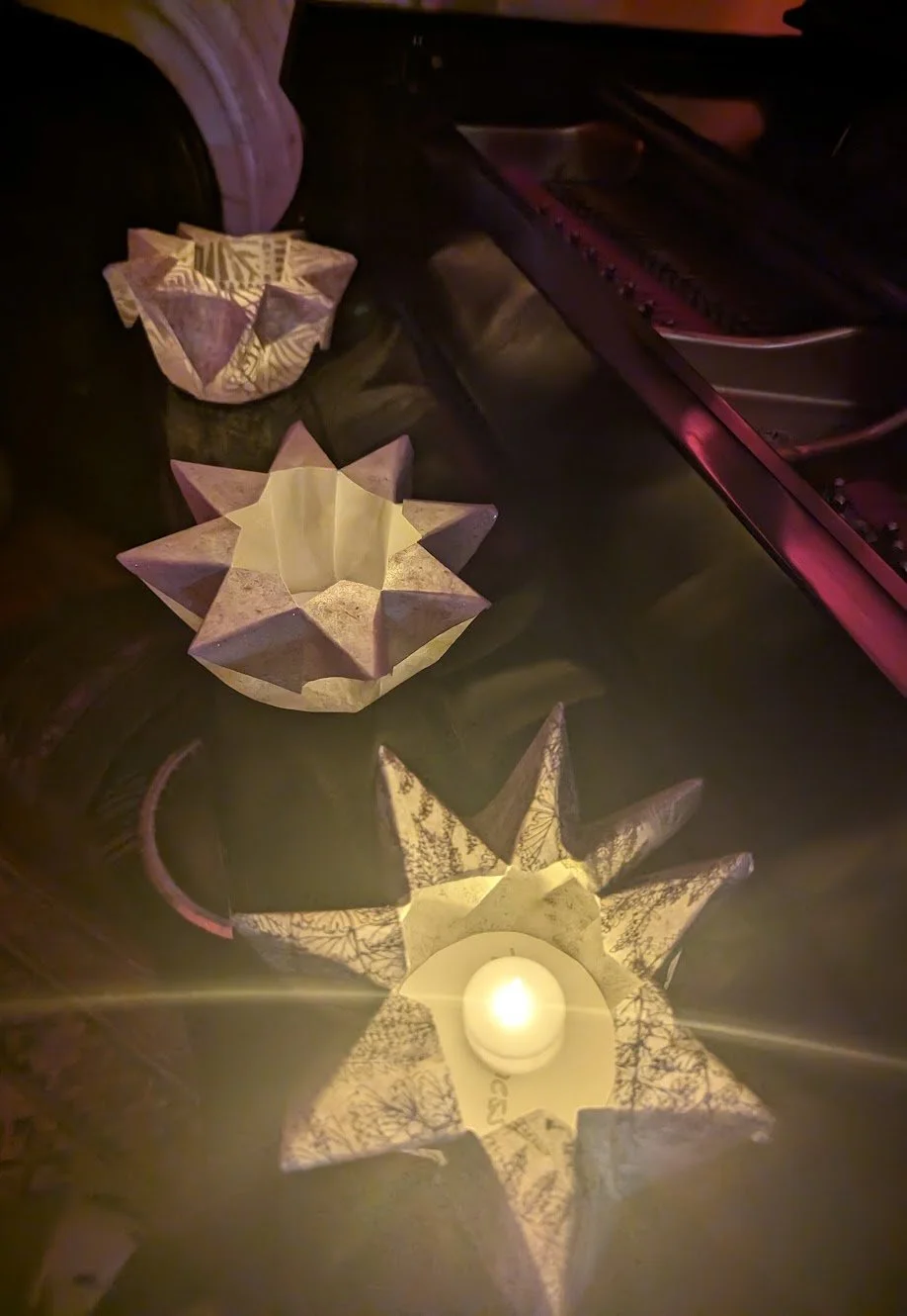 Three illuminated star-shaped paper lanterns with candles inside, placed on a reflective surface next to a dark-colored piano or keyboard.