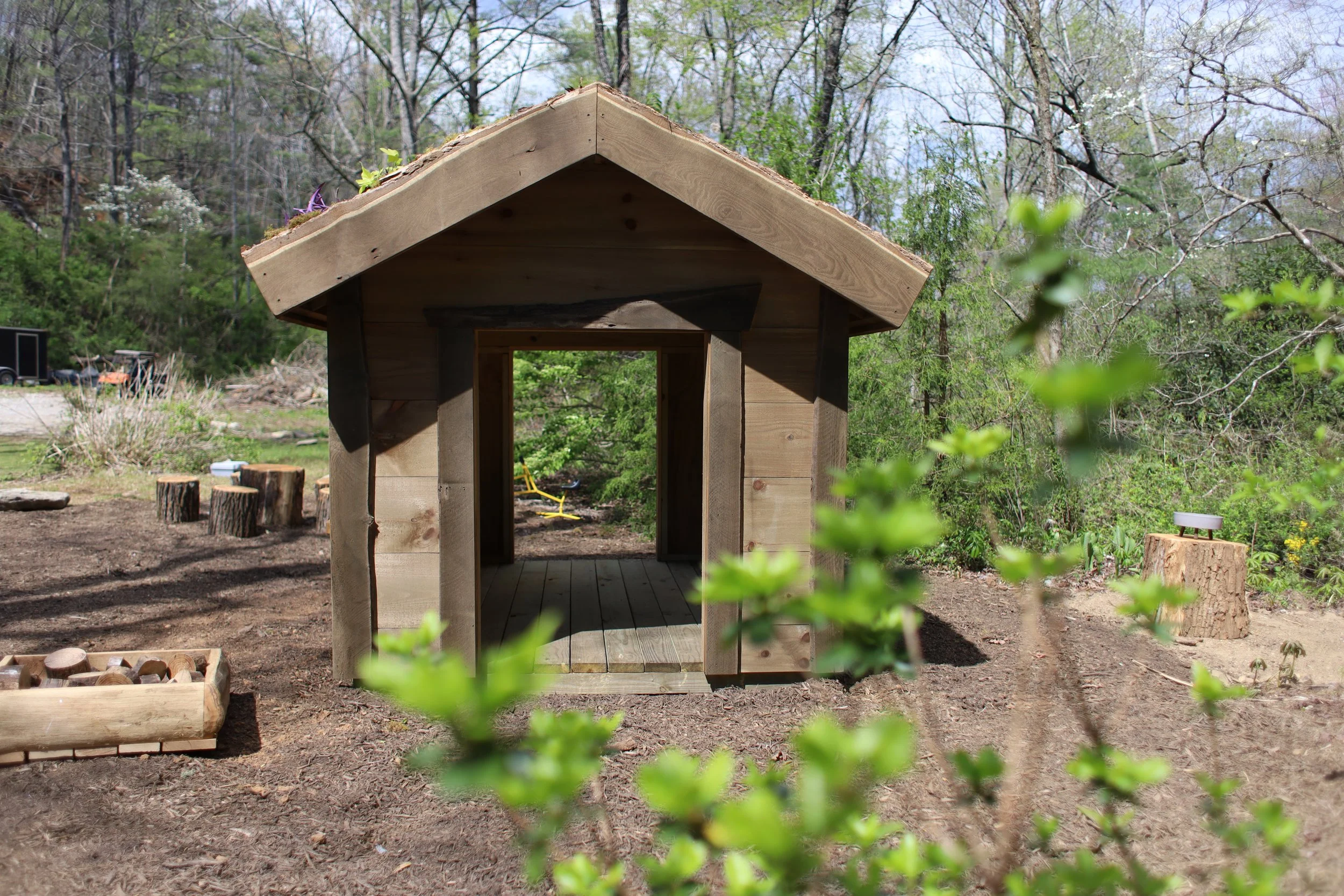 A small wooden structure, possibly a garden shed or playhouse, under construction in an outdoor setting with trees and greenery in the background.