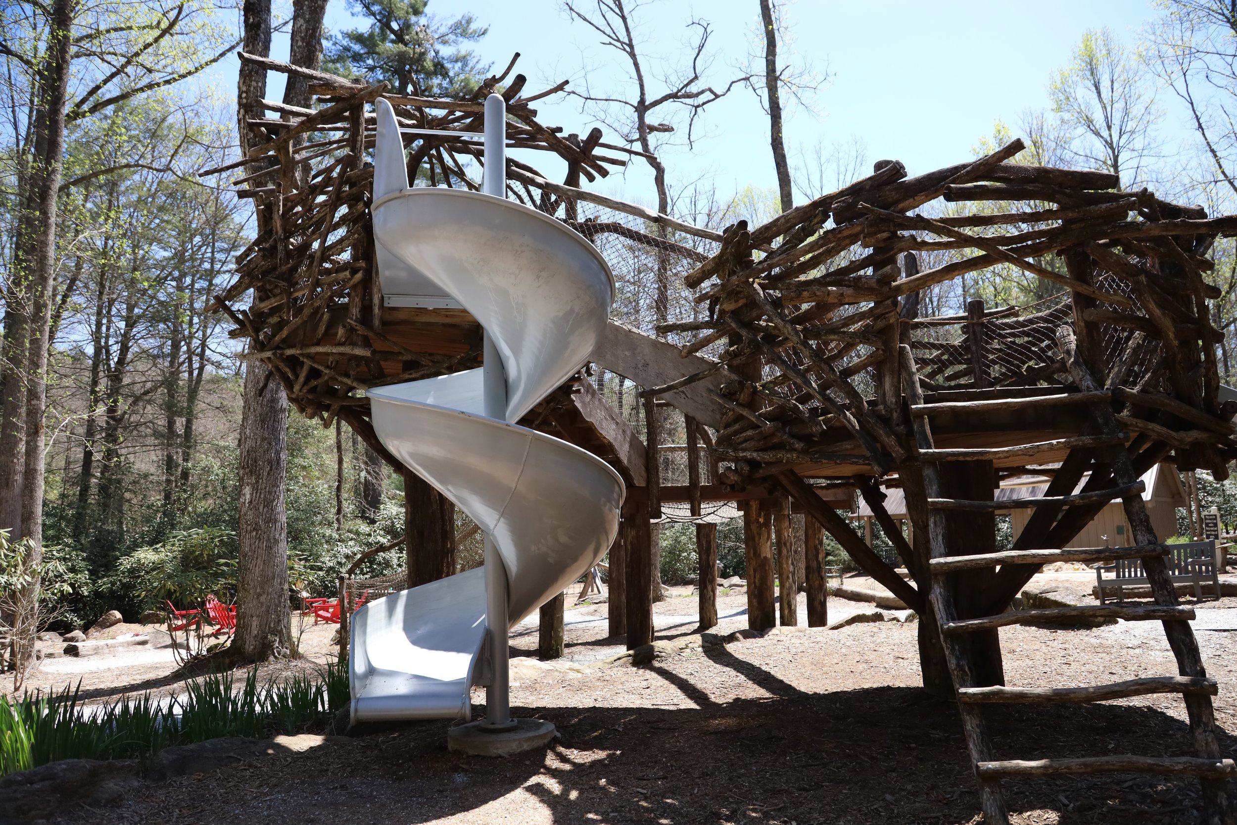 A wooden treehouse structure with a white spiral slide at a park surrounded by trees with some green foliage and a clear blue sky.