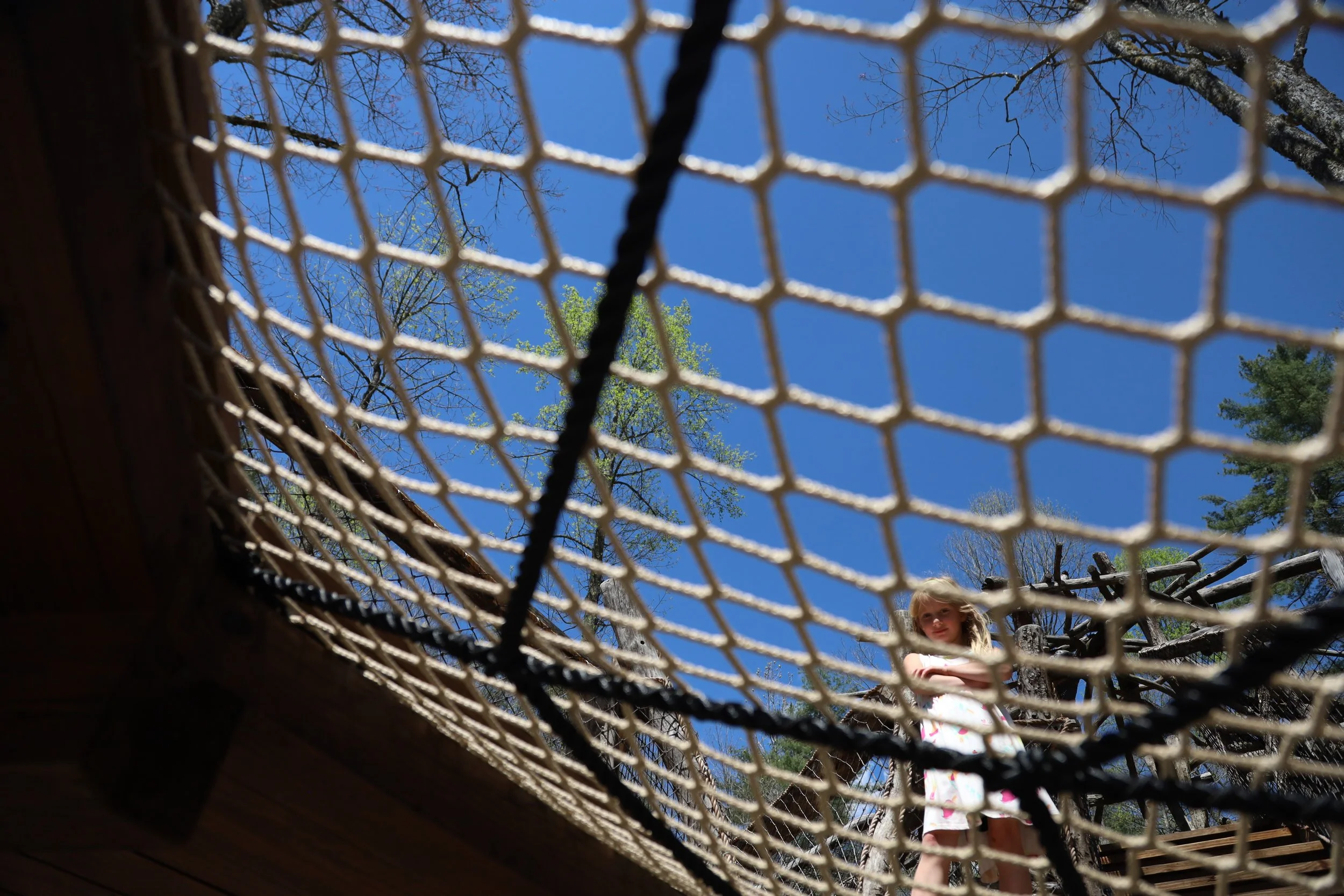 Looking up through a tennis net at a young girl standing outside on a sunny day with a blue sky and trees in the background.