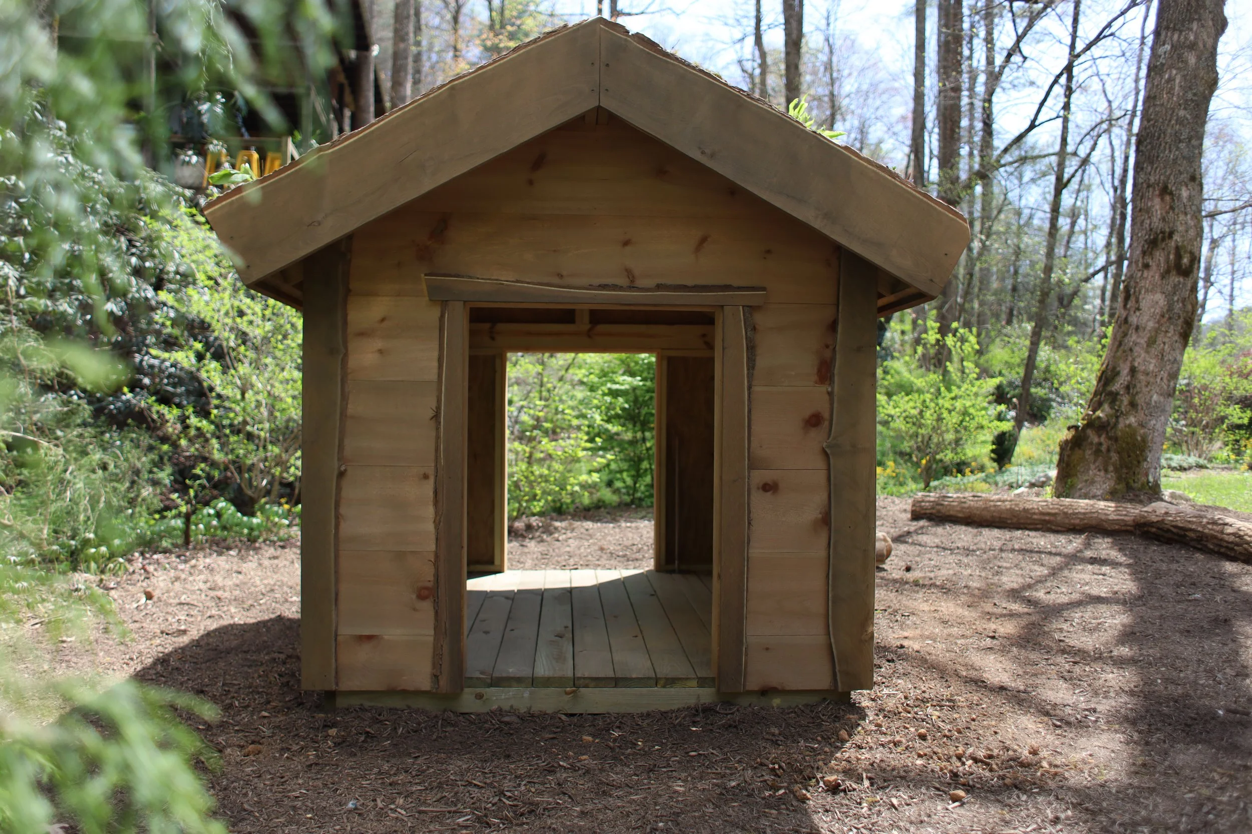 A small wooden playhouse with an open front door, situated outdoors in a forested area with trees and greenery surrounding it.