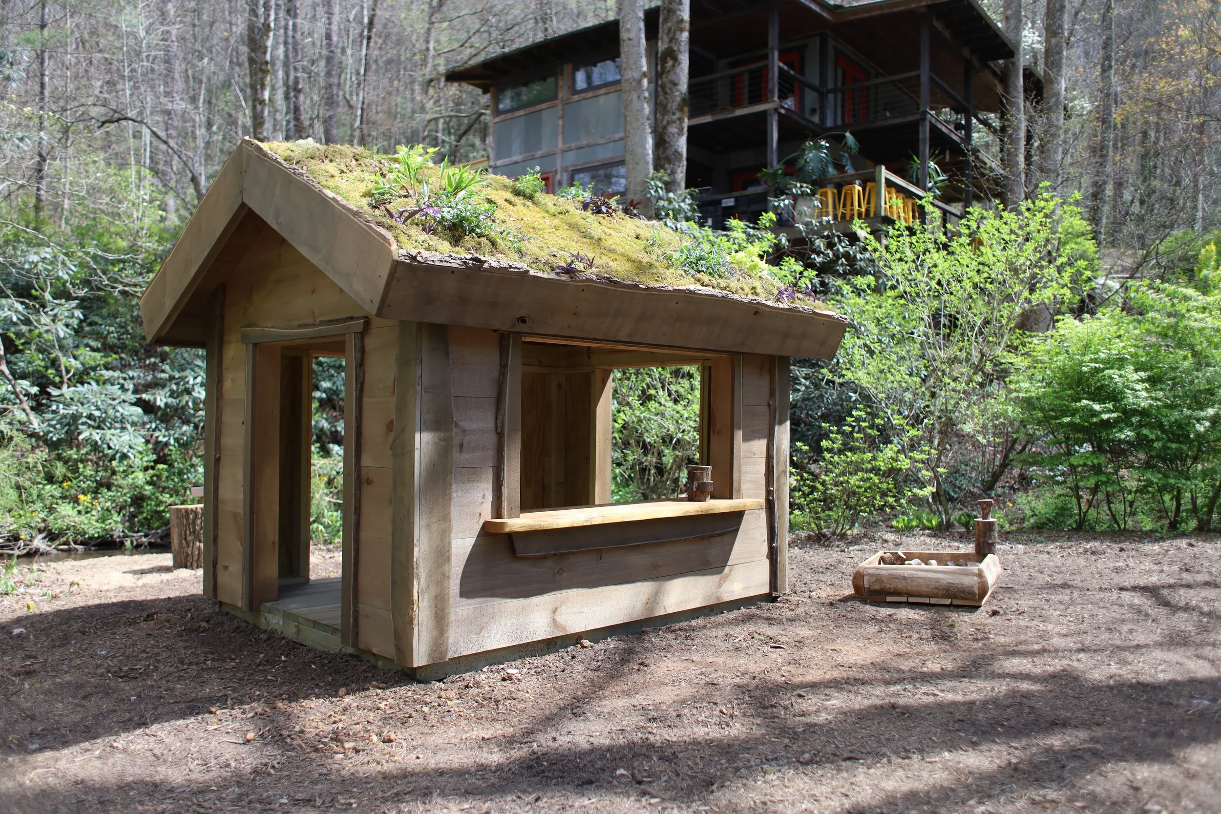 A small, unfinished wooden playhouse with a green moss-covered roof, located outdoors among trees and bushes, with a larger house visible in the background.