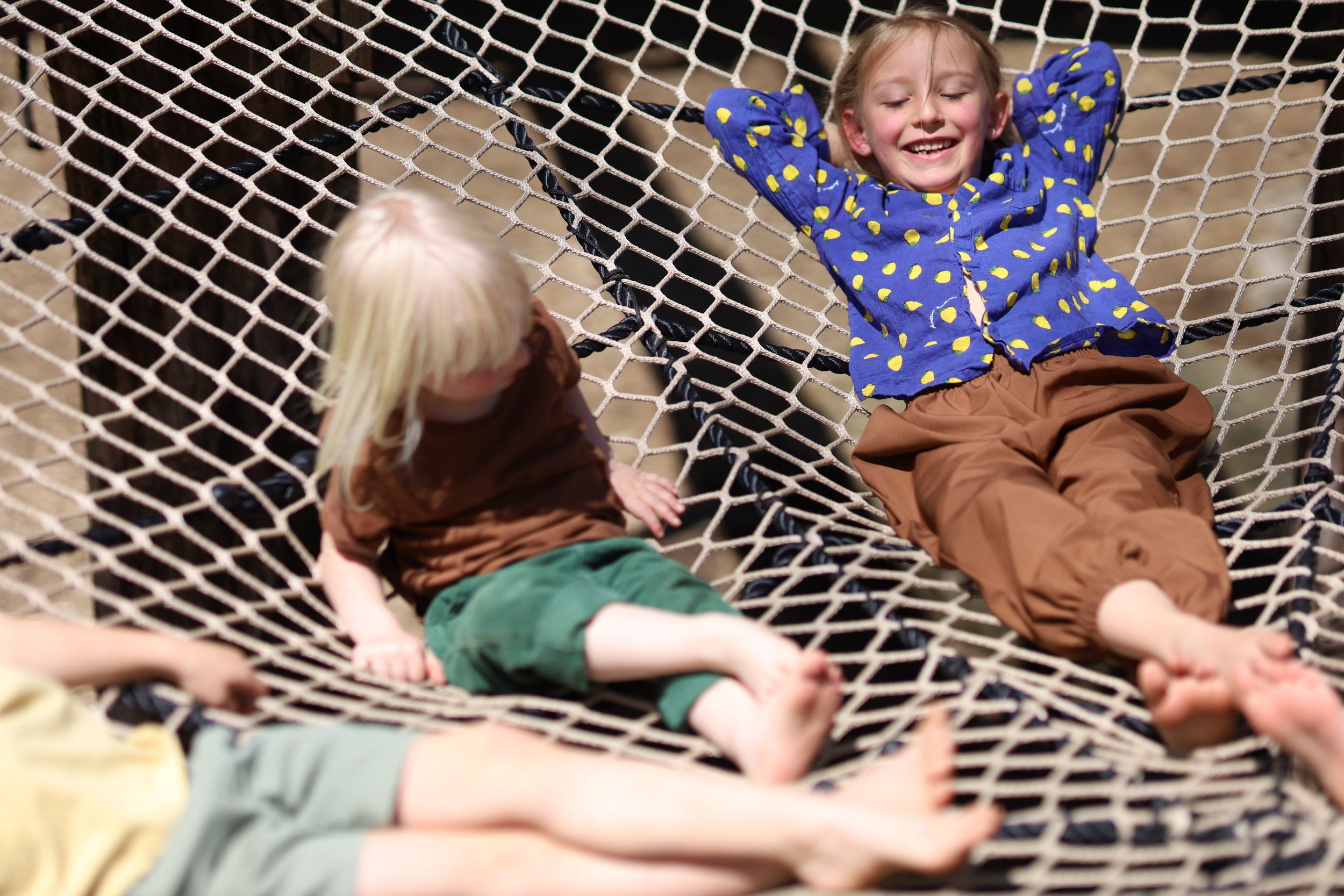 A young girl with a blurred face lying on a hammock with another child, both facing each other, outdoors.
