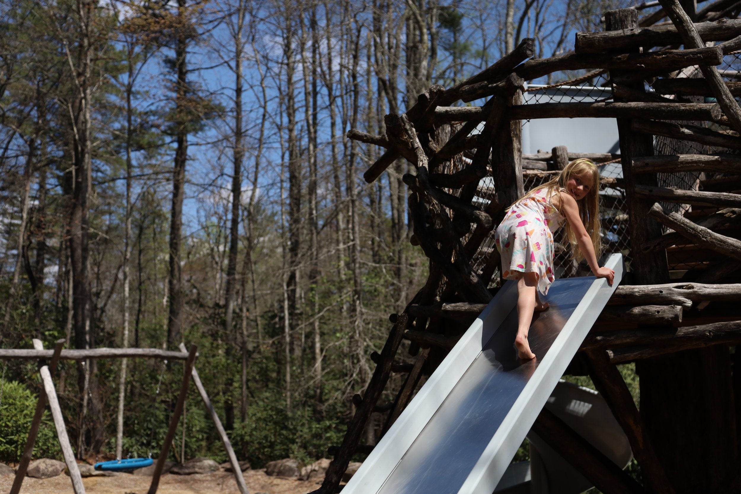 A young girl in a colorful dress with pink flamingos is climbing a slide at a playground surrounded by trees on a sunny day.