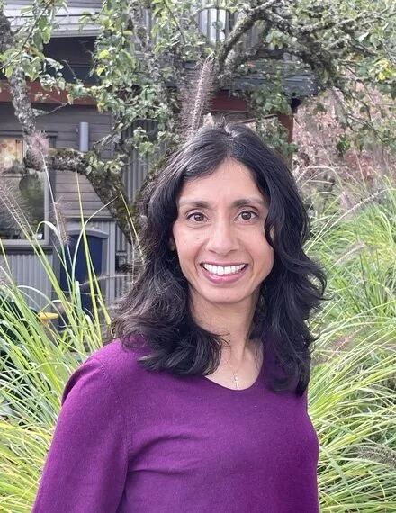 Woman with long dark hair wearing a purple top, smiling outdoors with trees and greenery in the background.