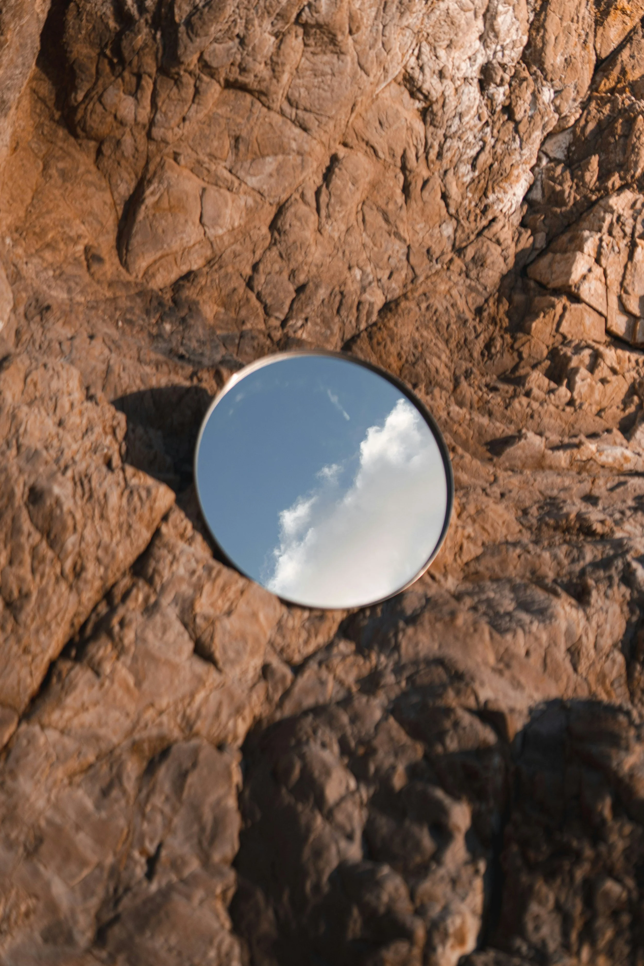 A round mirror placed on rough, reddish-brown rocky ground reflecting the blue sky with a few white clouds.