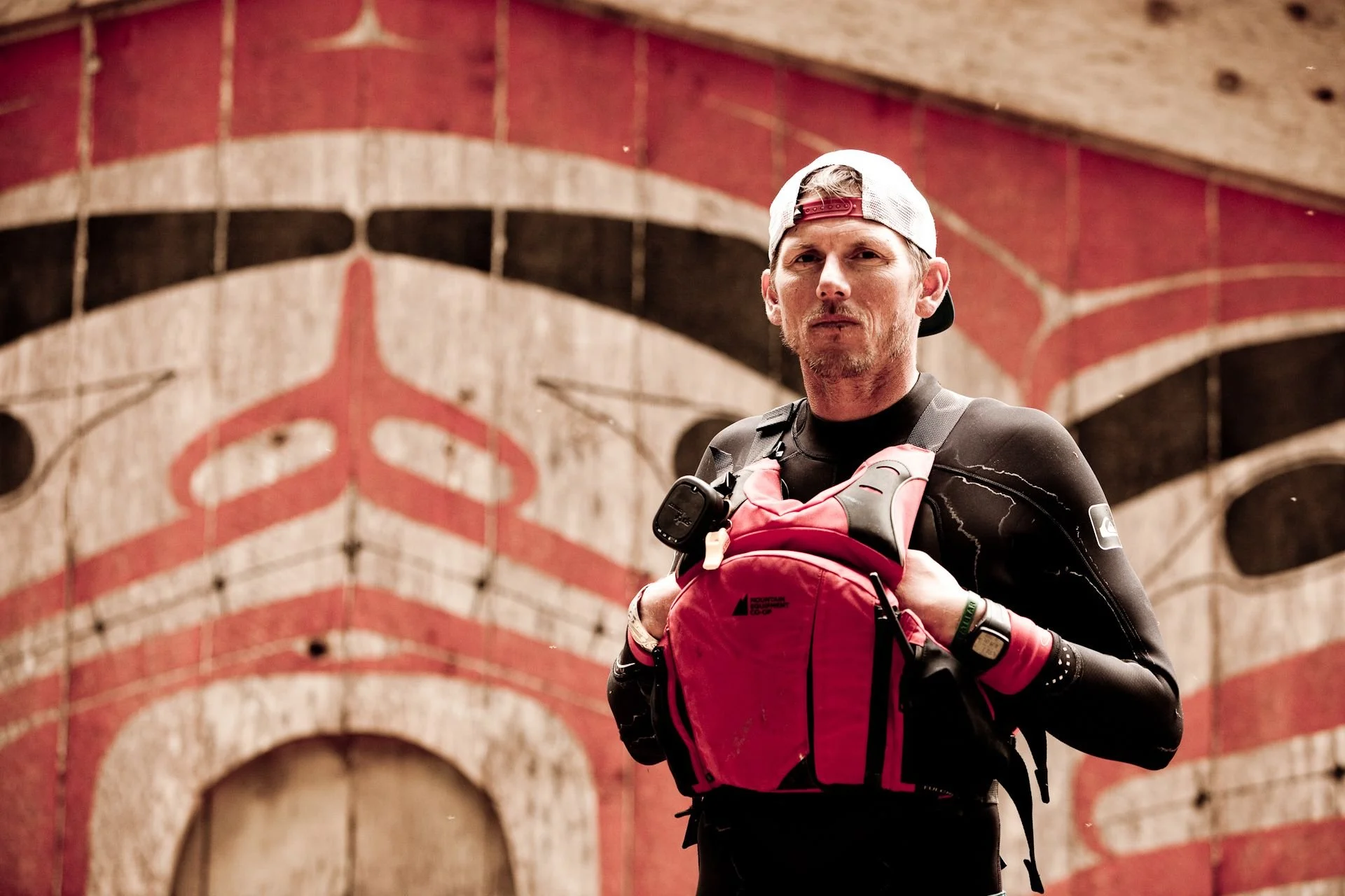 A man wearing a white and maroon cap, black athletic gear, and pink gloves running with a red backpack in an indoor archway with a painted red and black design.
