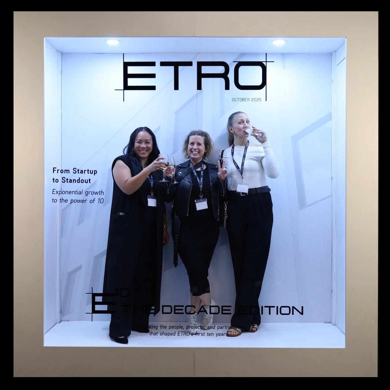 Three women standing together and smiling at an event, with a white backdrop featuring the text 'ETRO' and 'October 2025'. They are holding wine glasses and making celebratory gestures.