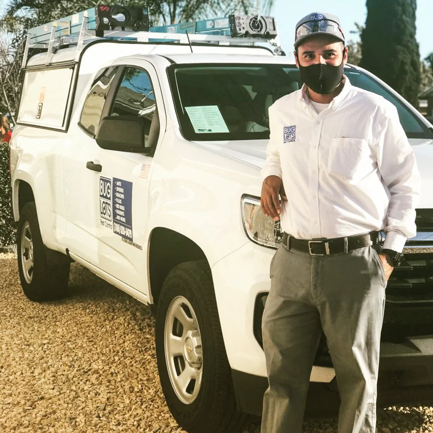 A man wearing a white shirt and gray pants standing next to a white service truck with a ladder rack on top, logo decals on the door, and a black face mask, outdoors on a gravel surface.