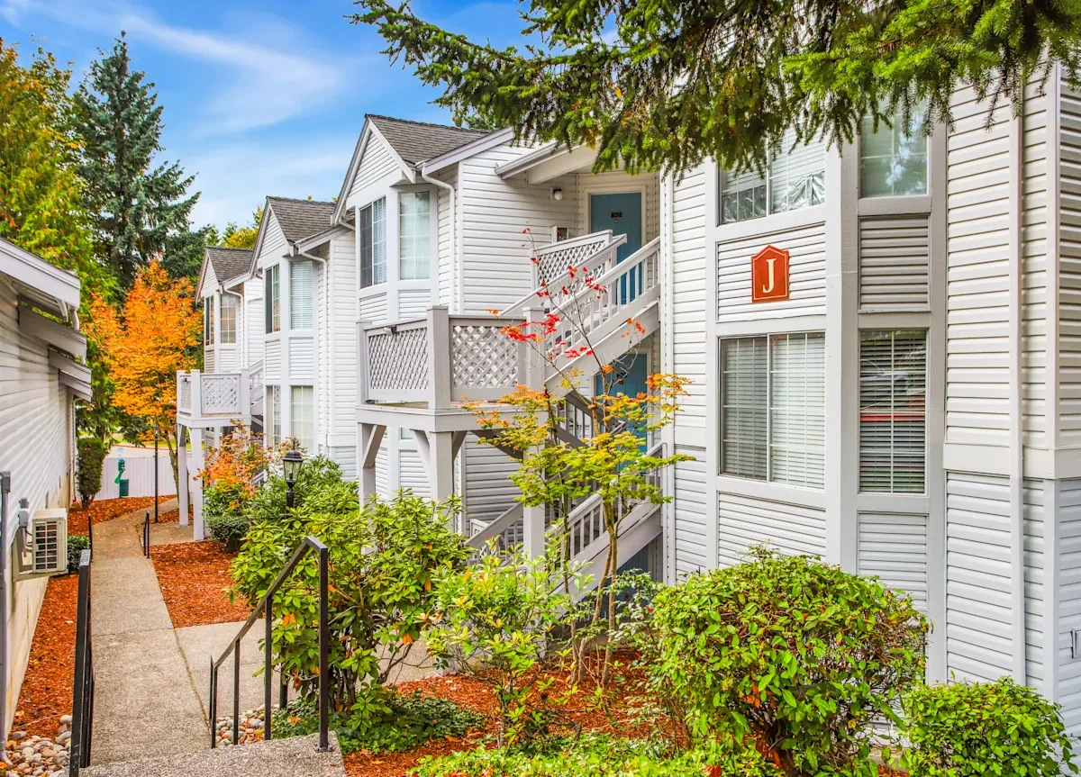 Exterior view of a white multi-family residential complex with balconies and stairs, surrounded by landscaped greenery and trees with fall foliage.