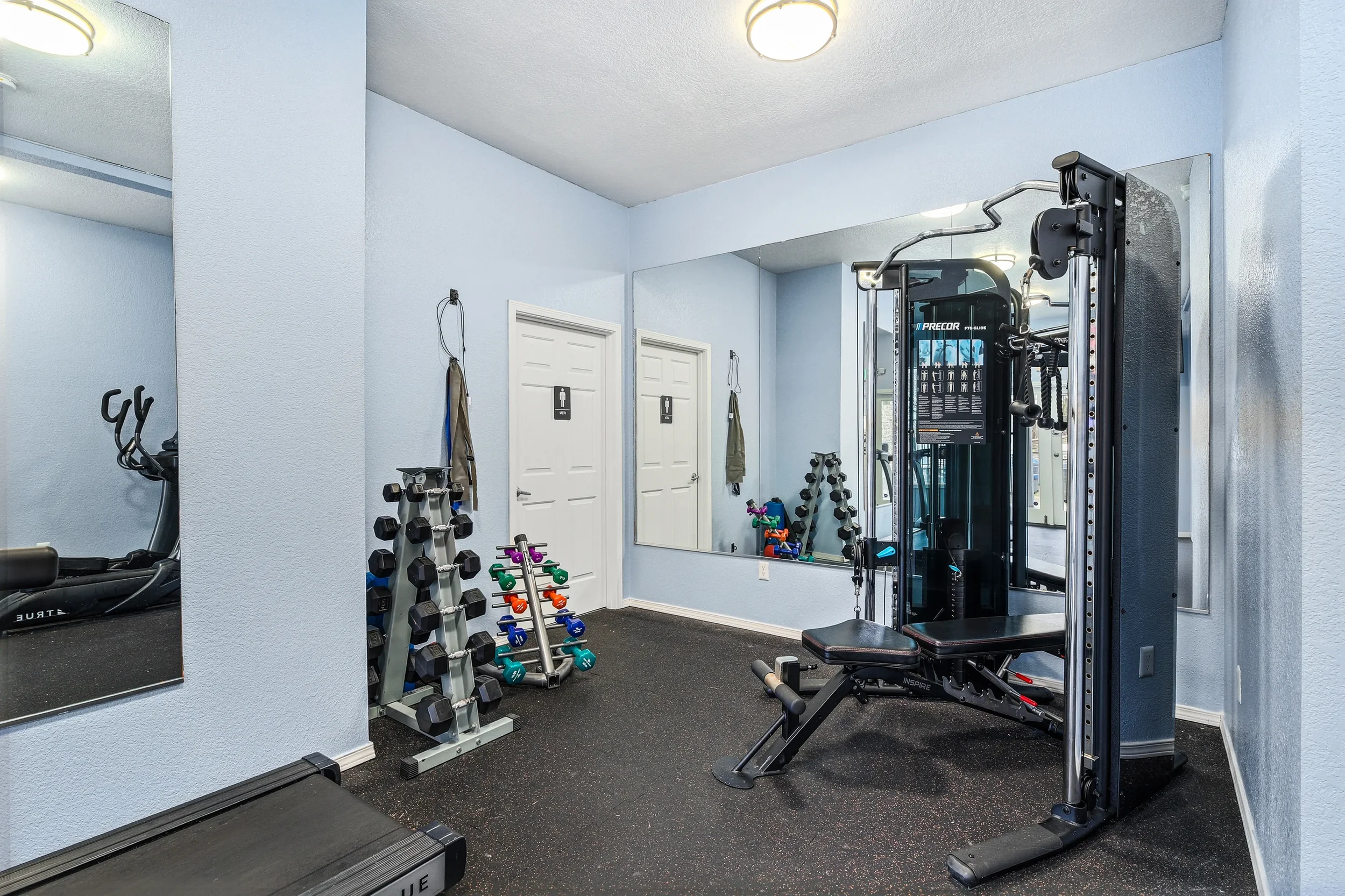 Home gym with light blue walls, black rubber flooring, and various workout equipment including dumbbells, resistance bands, a weight machine, and a treadmill.
