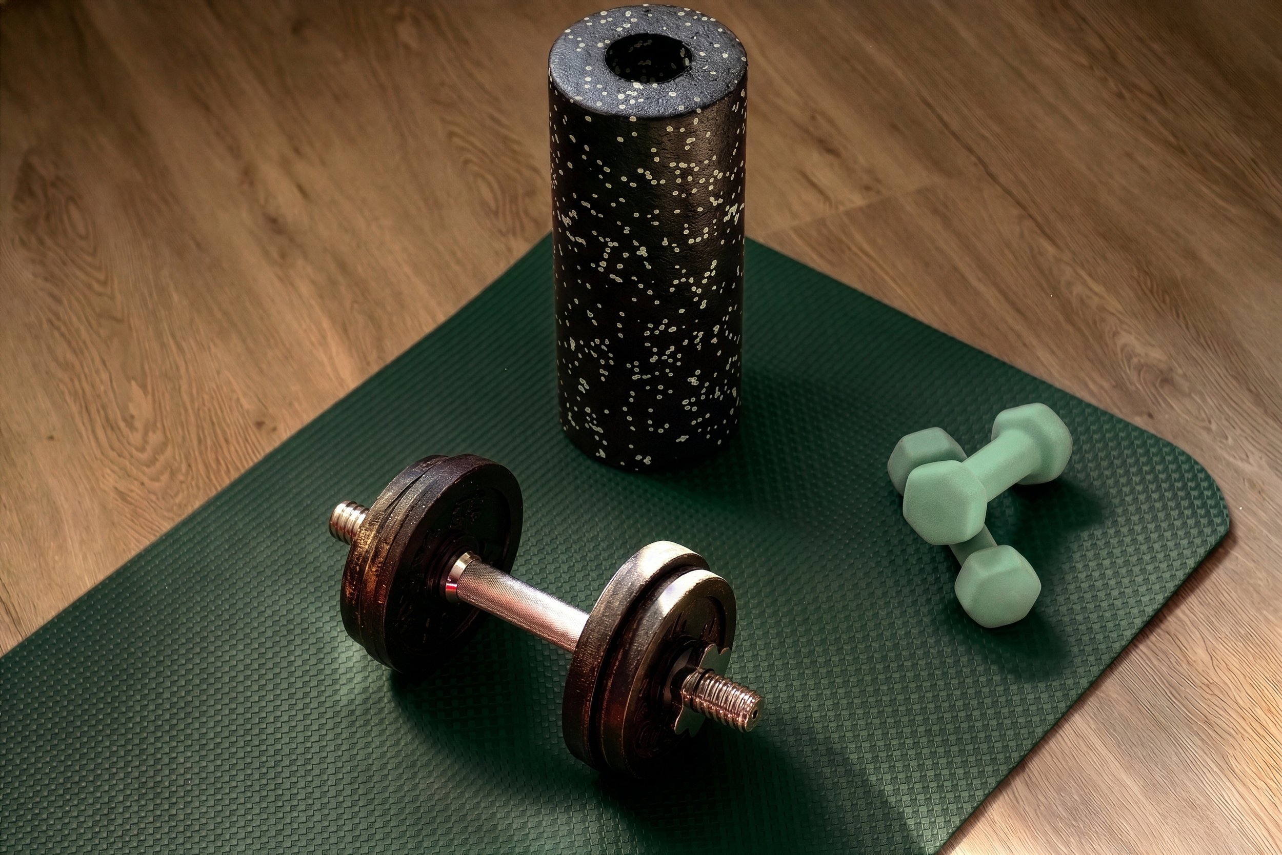 A workout area with a green exercise mat on wooden floor, featuring dumbbells, a foam roller, and a pair of light green dumbbells.