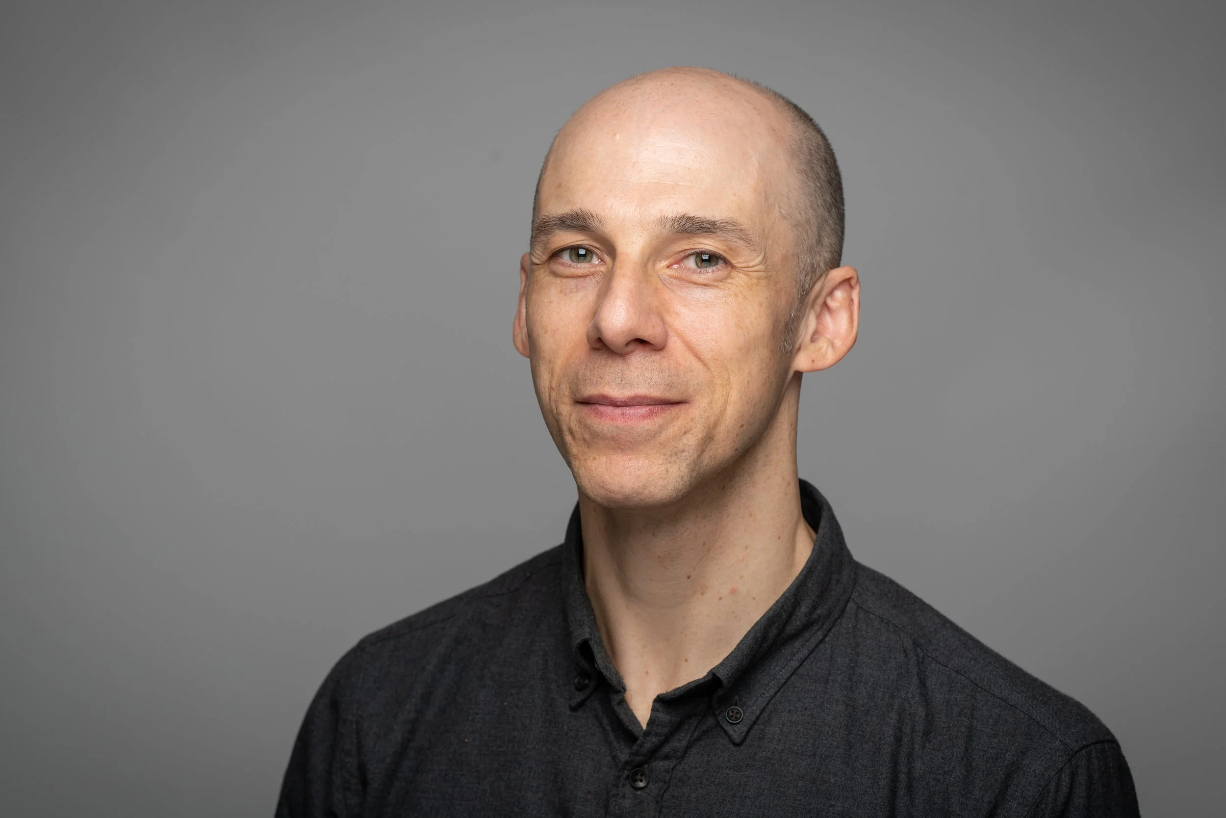 Portrait of a bald man with light skin and green eyes, wearing a dark gray collared shirt, against a plain gray background.
