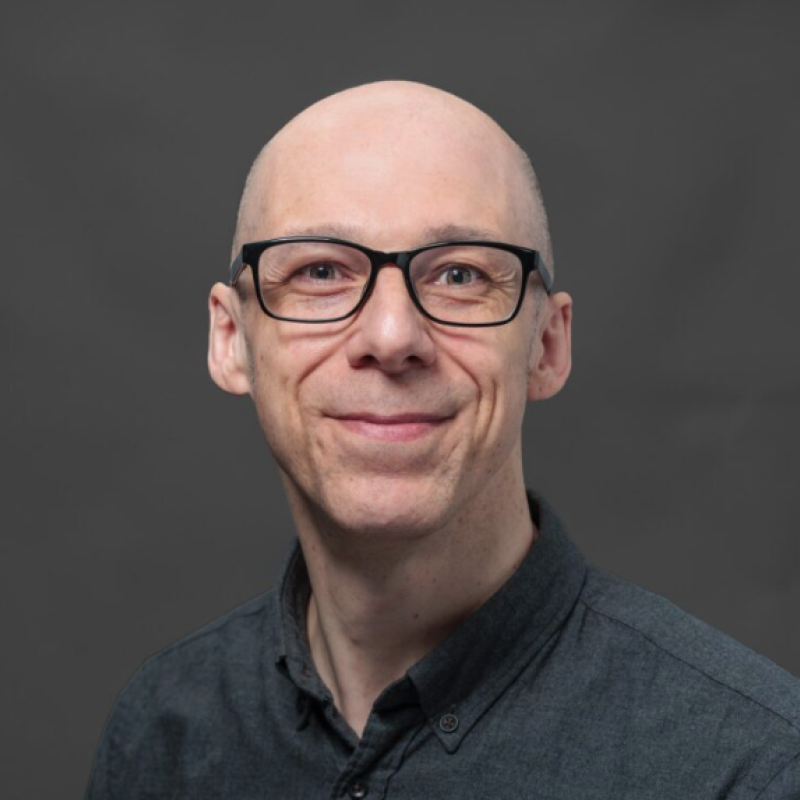 Headshot of a smiling man with glasses, wearing a dark button-up shirt, against a gray background.