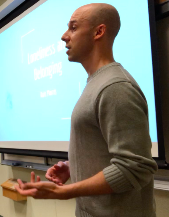 A man giving a presentation in a classroom, standing in front of a large screen with a blue slide titled 'Loneliness and Belonging.'