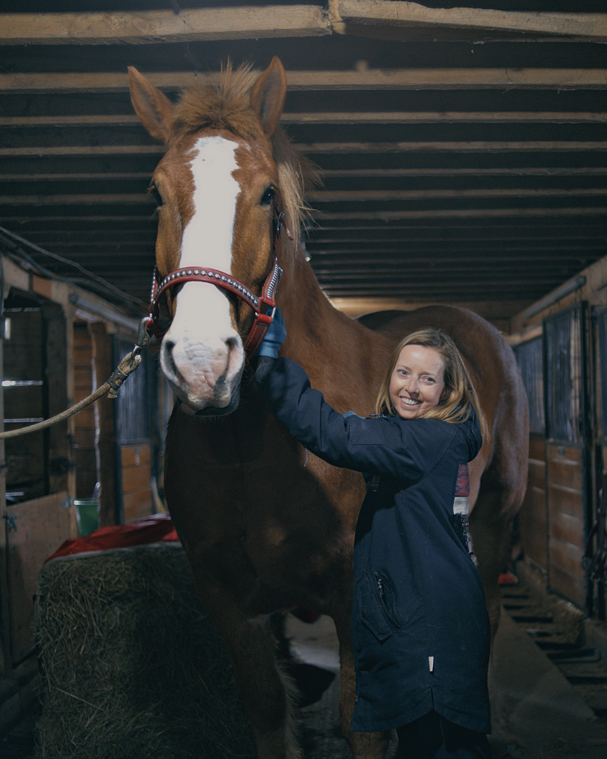 A woman hugging a brown horse with a white stripe down its face inside a stable.