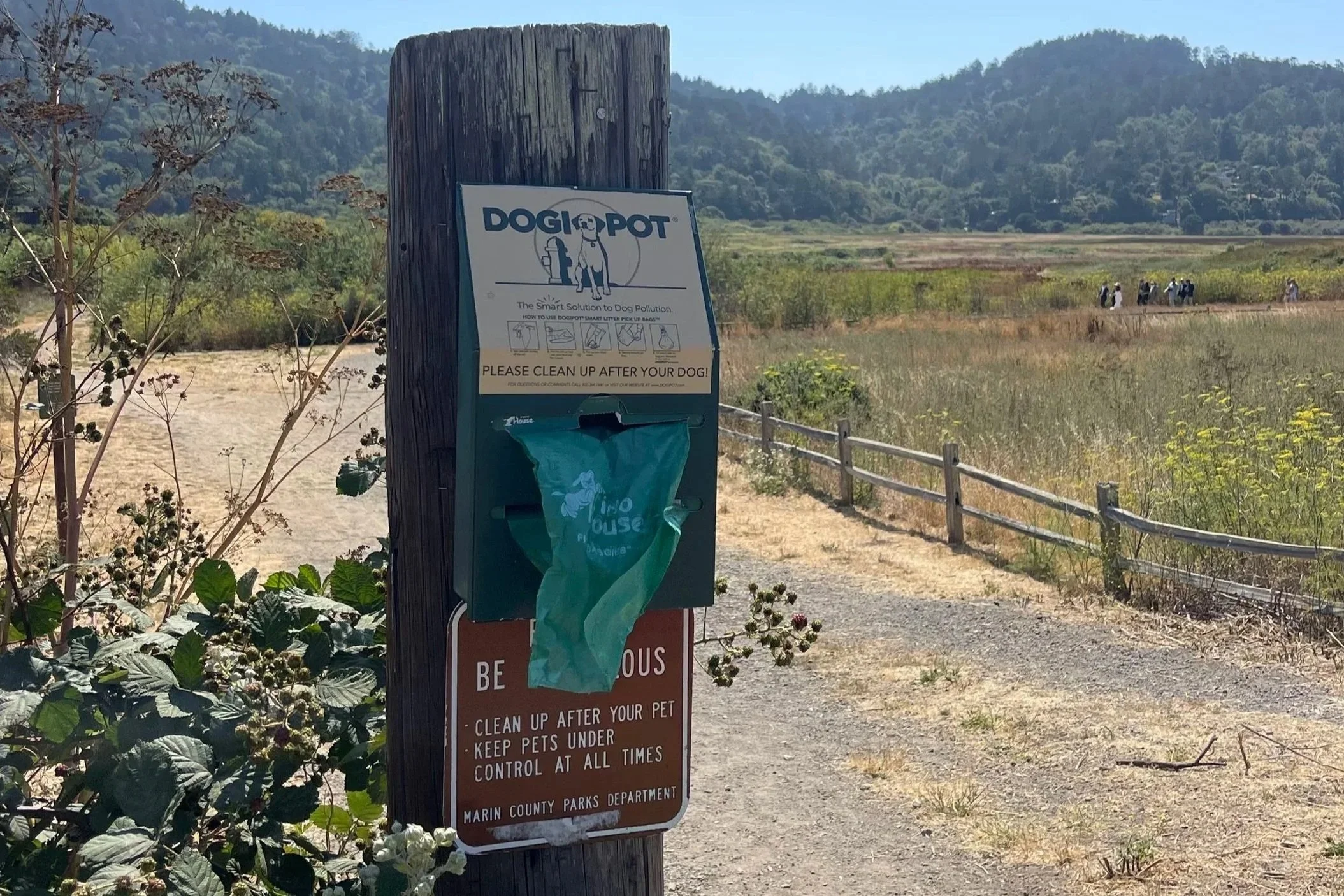 Sign on a post with instructions for dog owners at a park. In the background, there is a mountain landscape with some people walking on a trail.
