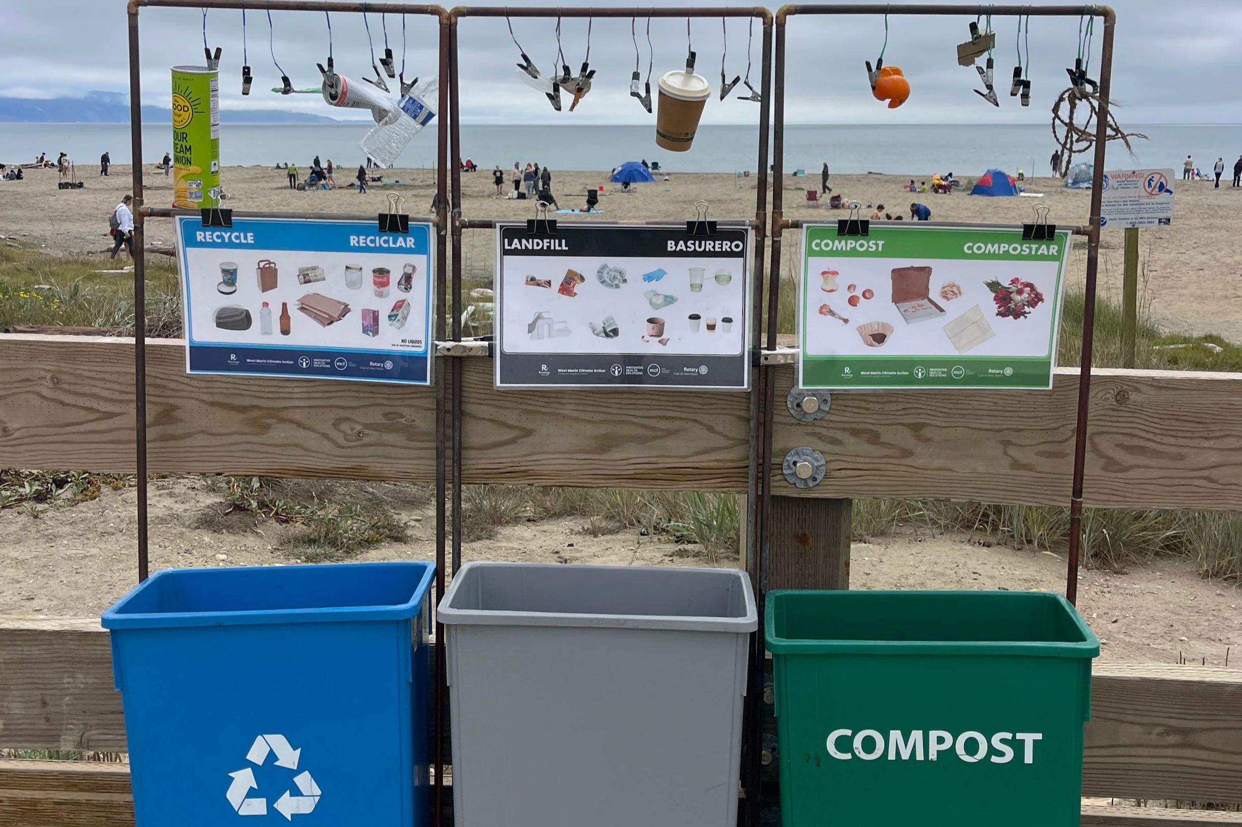 Recycling and compost bins at the beach with informational signs showing types of waste appropriate for each bin.