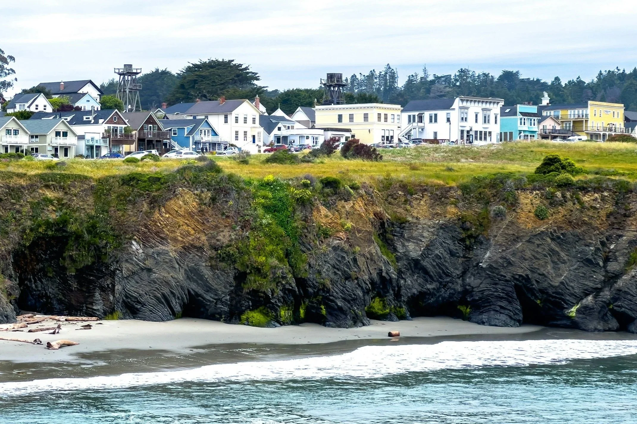 Colorful houses with water towers on a hill overlooking the ocean with rocky cliffs in the foreground and trees in the background.