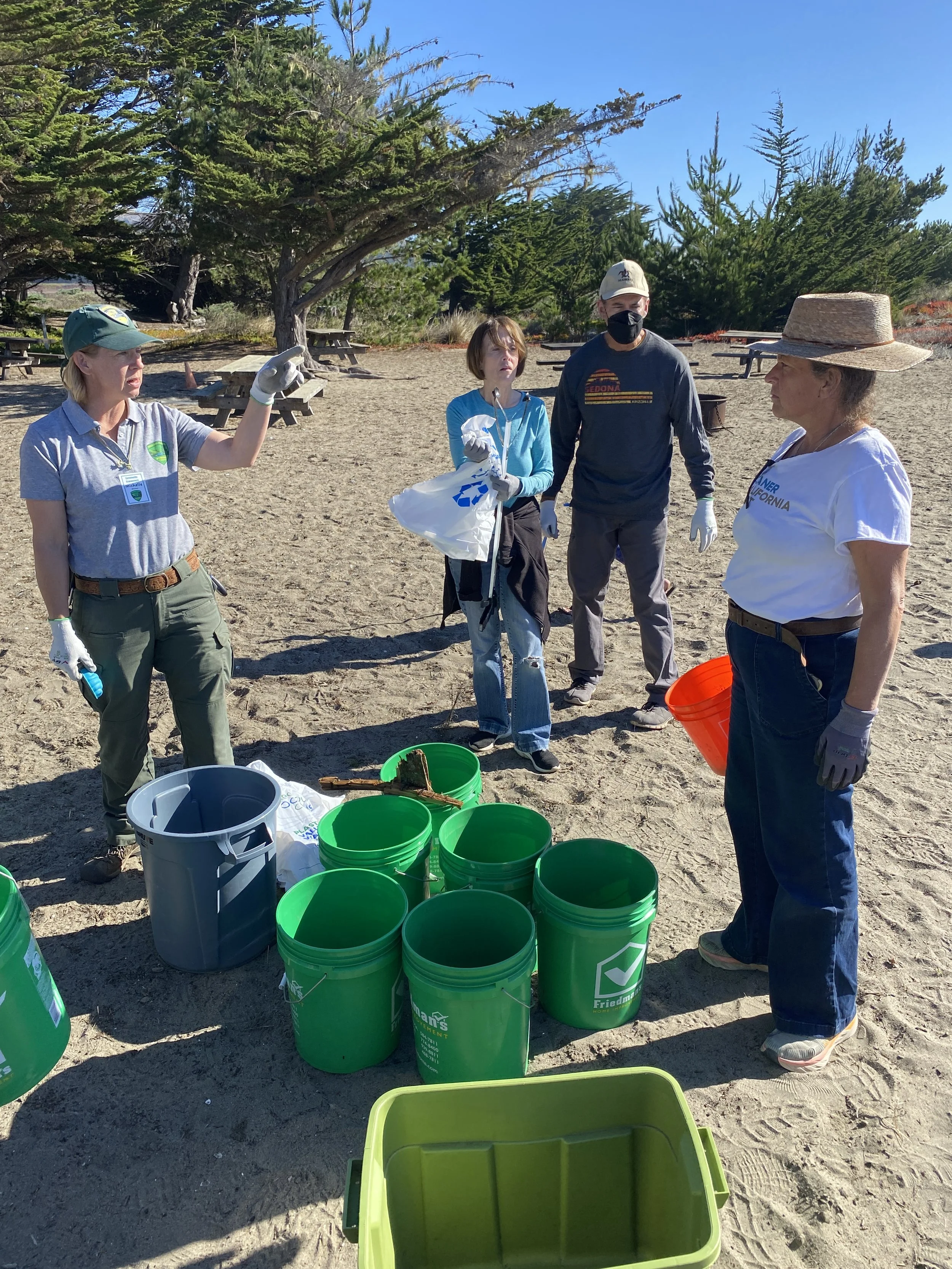 Four people standing outdoors near trees, with buckets and containers in front of them, apparently participating in a cleanup or volunteer activity on a sunny day.