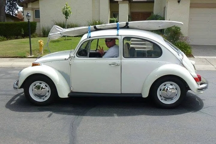 A white vintage Volkswagen Beetle car on the street with a surfboard attached on top. There is a person inside the car holding up two fingers, making a peace sign. The background shows a residential neighborhood with houses, a lawn, and a lamppost.