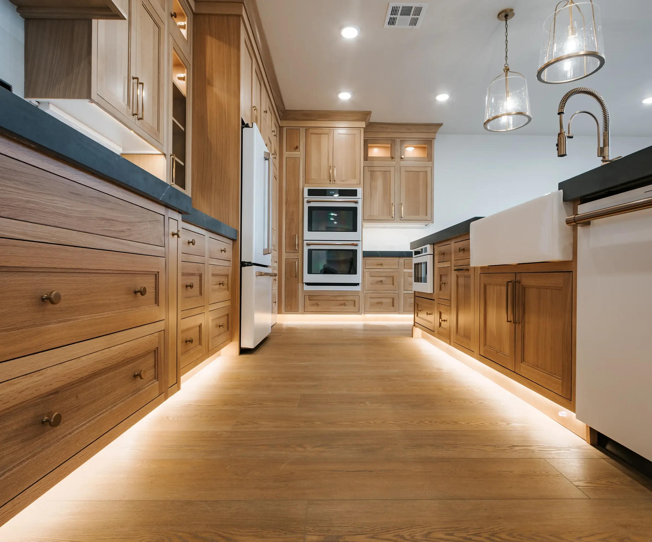 A modern kitchen with wooden cabinetry, black countertops, and built-in double ovens. Recessed lighting and pendant lights hang from the ceiling, and the flooring is wood.