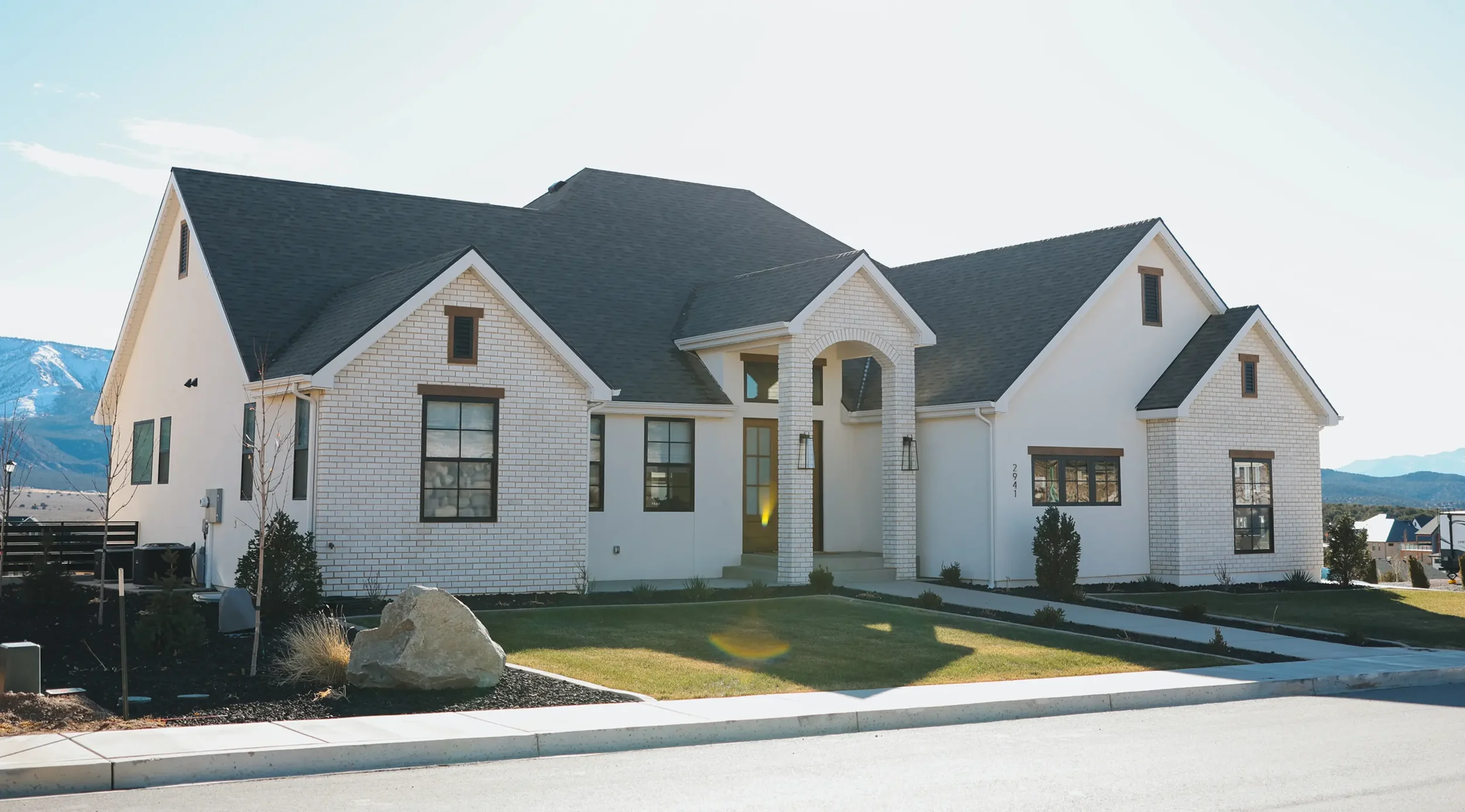 A modern two-story house with white brick exterior, black window frames, and a dark gray steeply pitched roof. The front yard has some young trees, bushes, and a large rock, with a sidewalk and street in front.