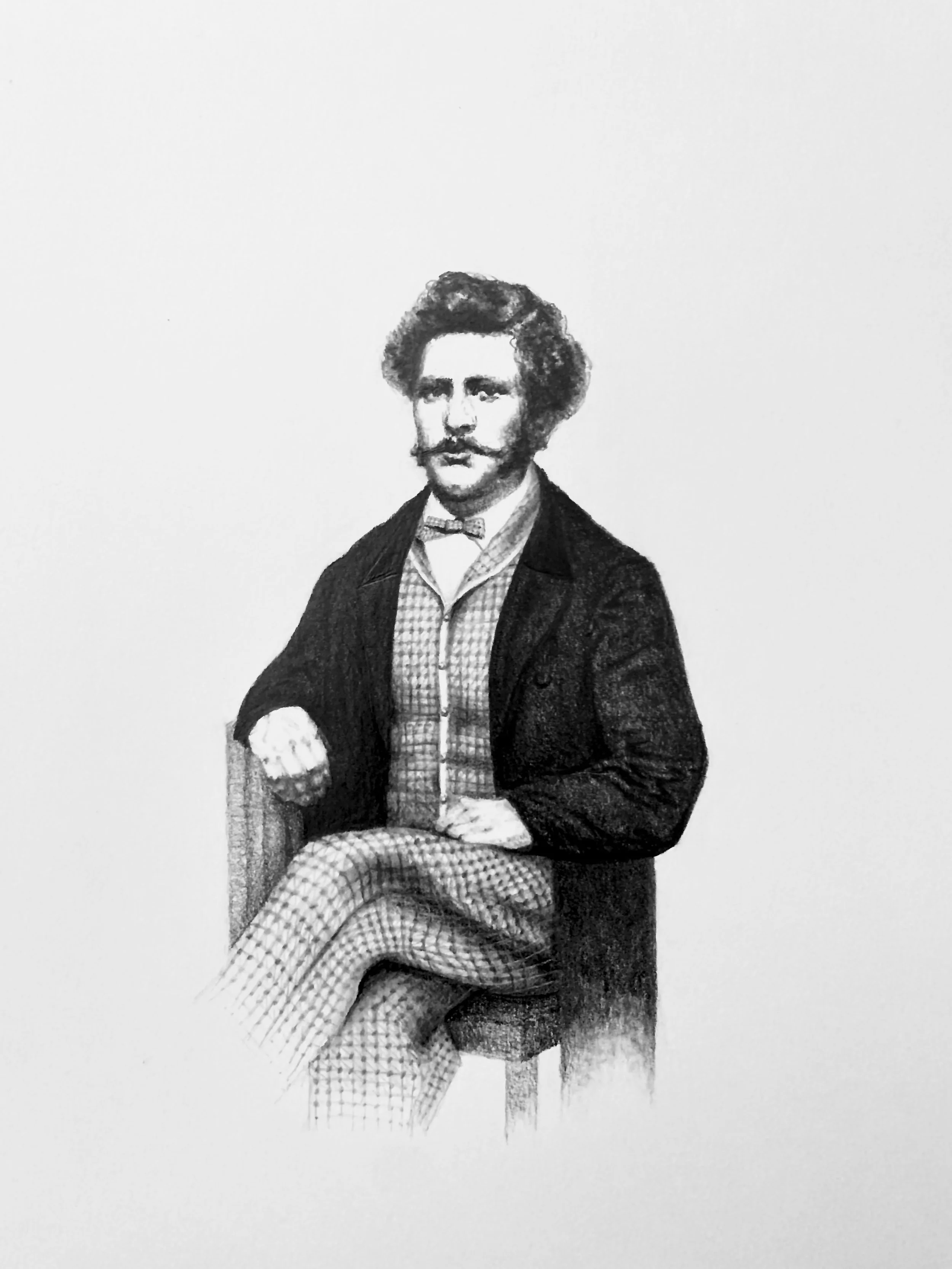 Black and white photograph of a man with curly hair, mustache, and glasses, dressed in vintage attire, seated across an empty background.