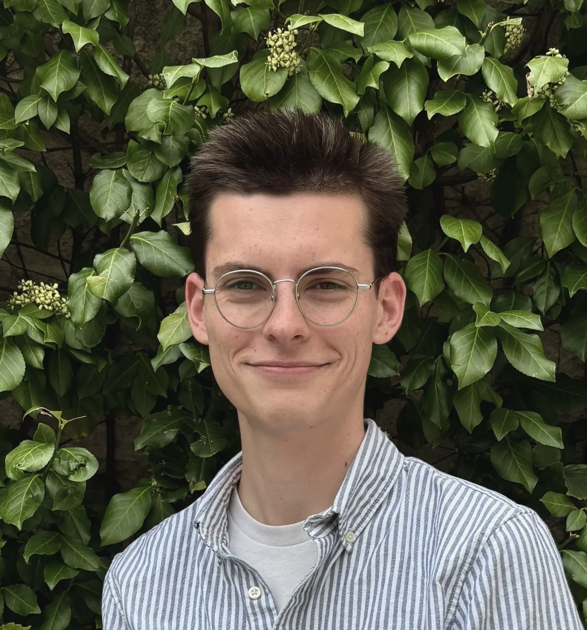 A young man with dark hair, wearing glasses and a striped shirt, smiling at the camera. He is standing in front of a hedge with large green leaves.