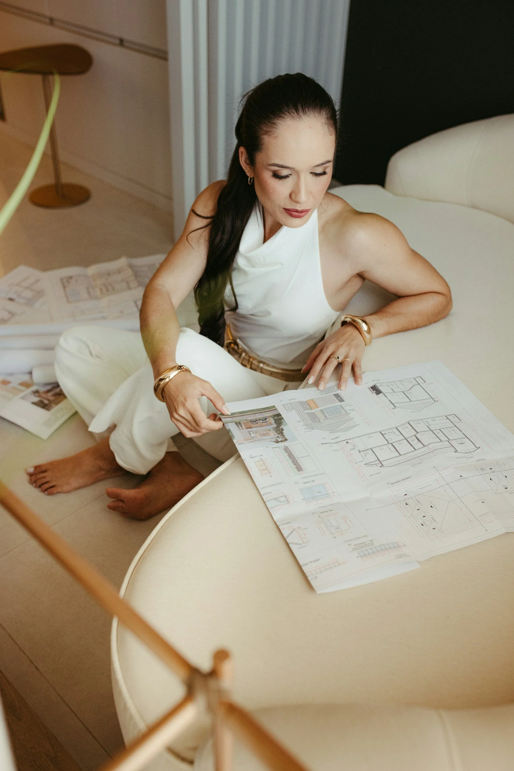 A woman sitting on the floor reviewing architectural blueprints, with additional drawings scattered around her. She has dark hair tied back, wears a sleeveless white top, and gold bracelets.