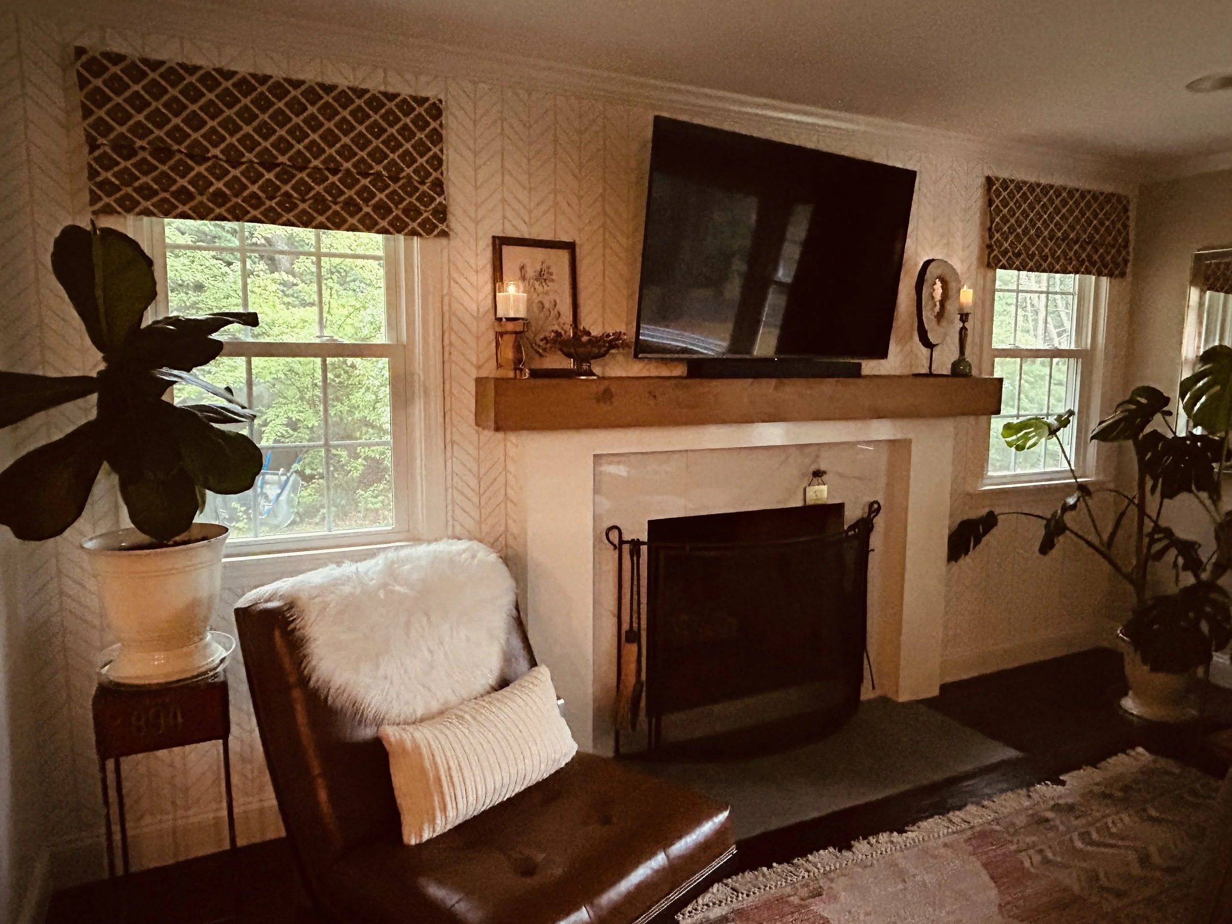 A living room with two windows, a mantle, television, brown leather chair with fur throw and pillows and plants
