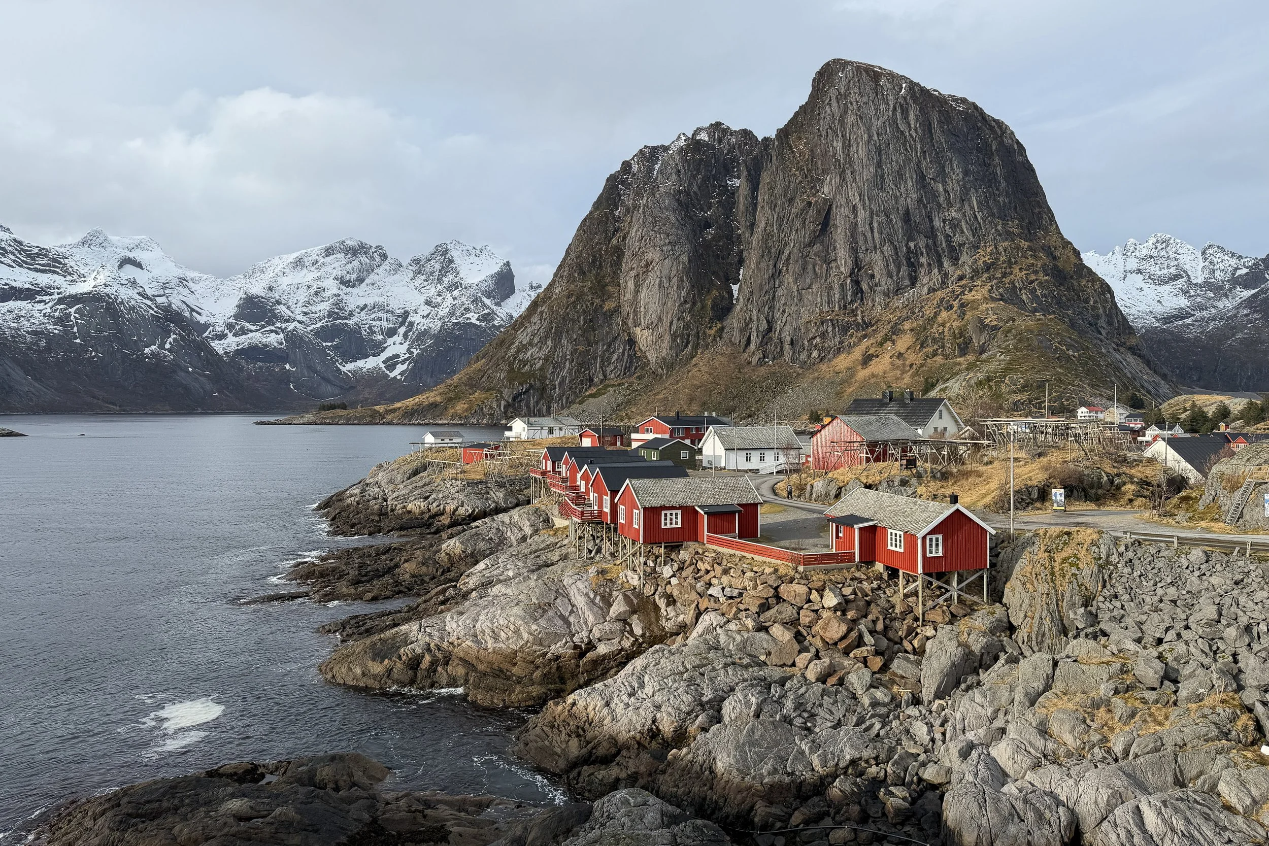 A scenic coastal village with red and white houses built on rocky shoreline, surrounded by snow-capped mountains and a calm body of water under a cloudy sky.