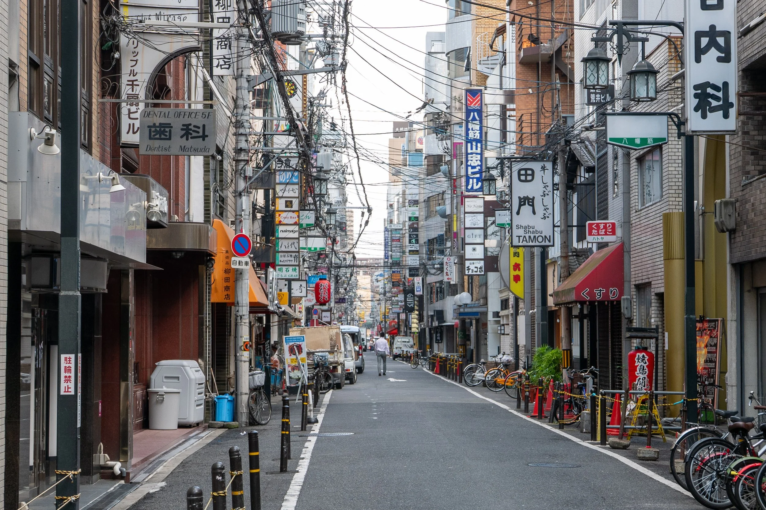 A narrow urban street in Japan filled with numerous signs in Japanese, bicycles, and a few pedestrians under a tangled network of overhead wires.