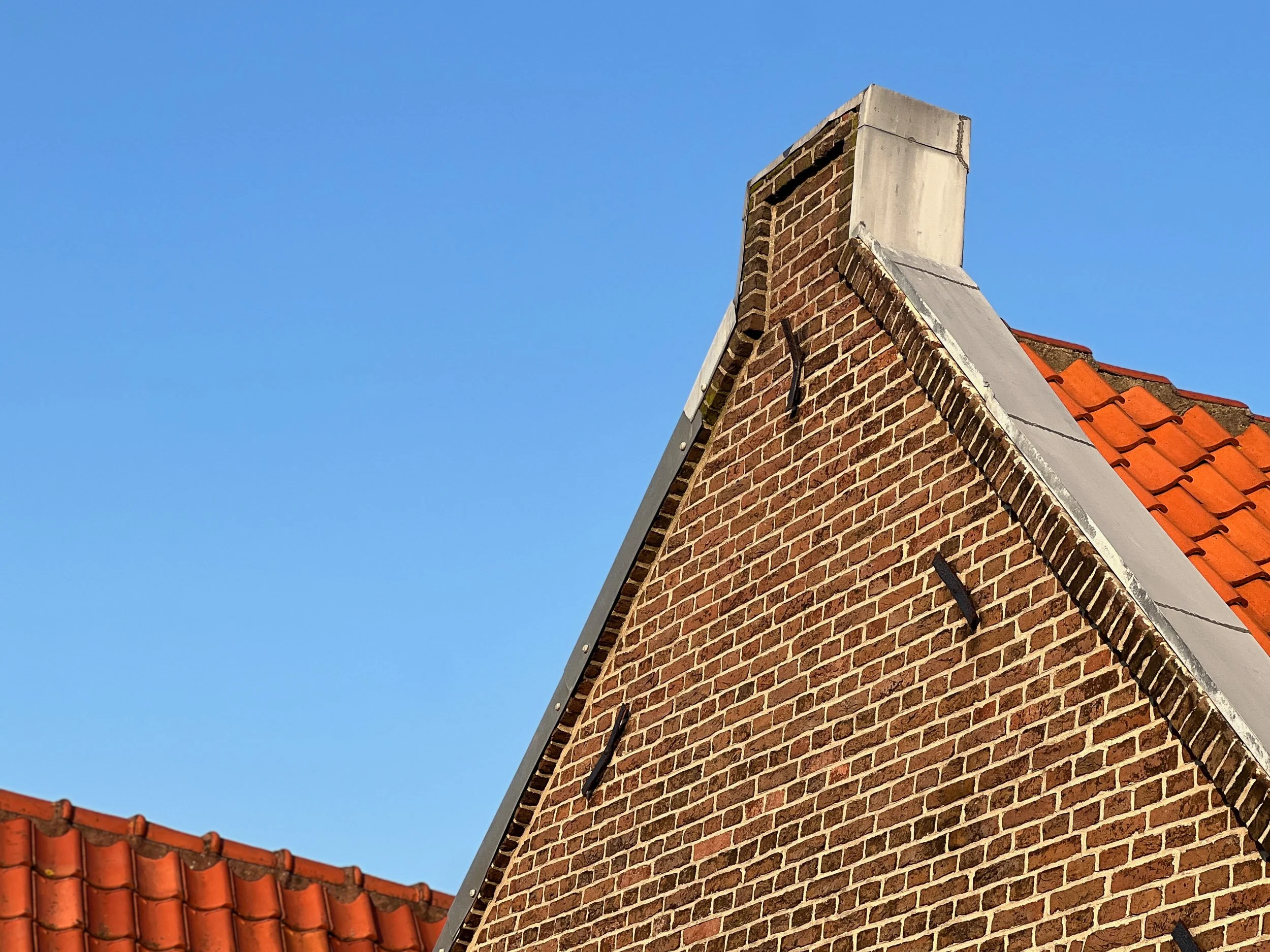 Close-up of a brick chimney with a concrete cap on a house roof, against a clear blue sky with orange-red roof tiles visible at the bottom.