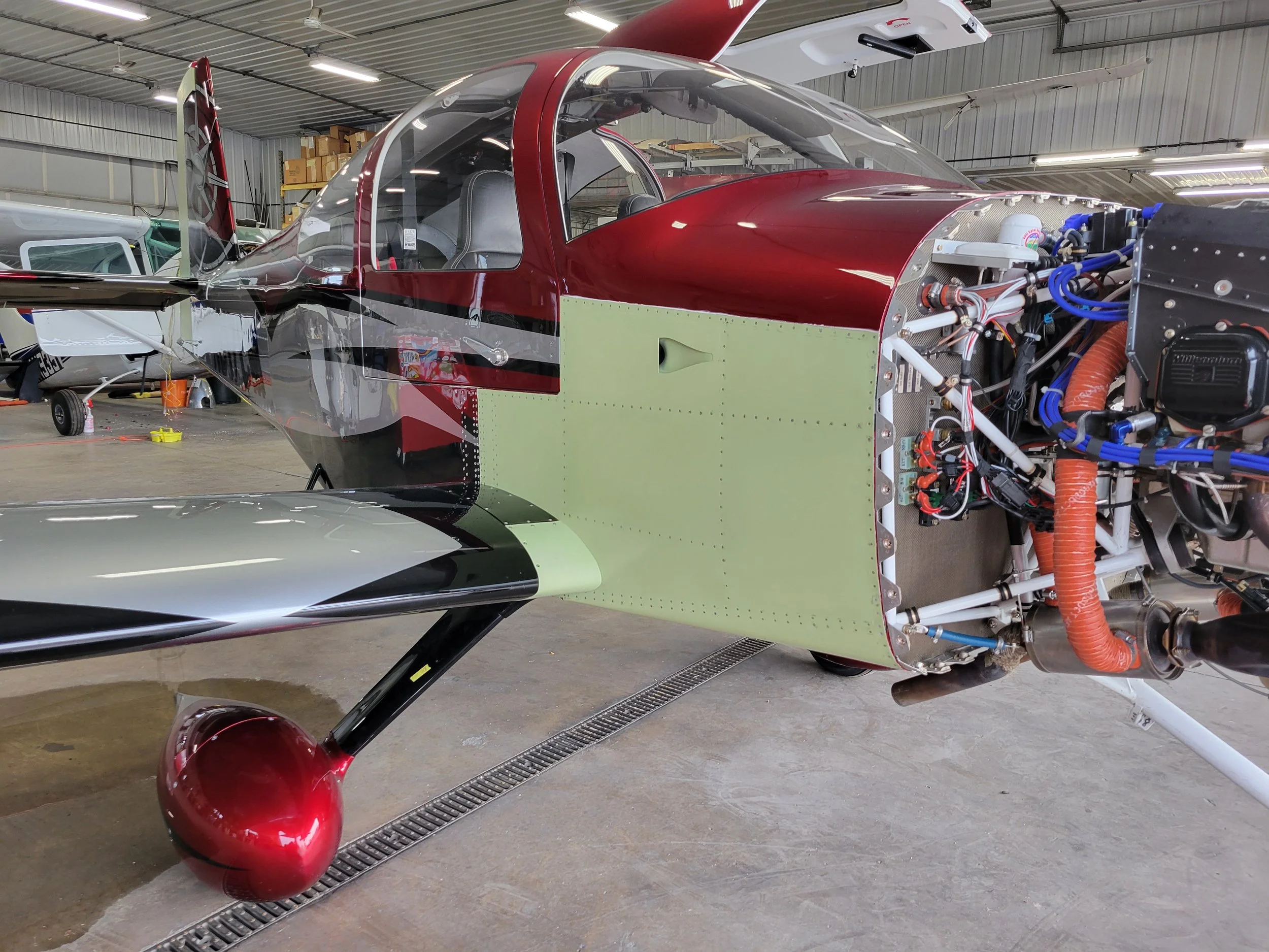 The image shows a partially assembled aircraft inside a hangar. The aircraft has a red and black exterior with a clear canopy, and there are visible internal components and wiring at the front.