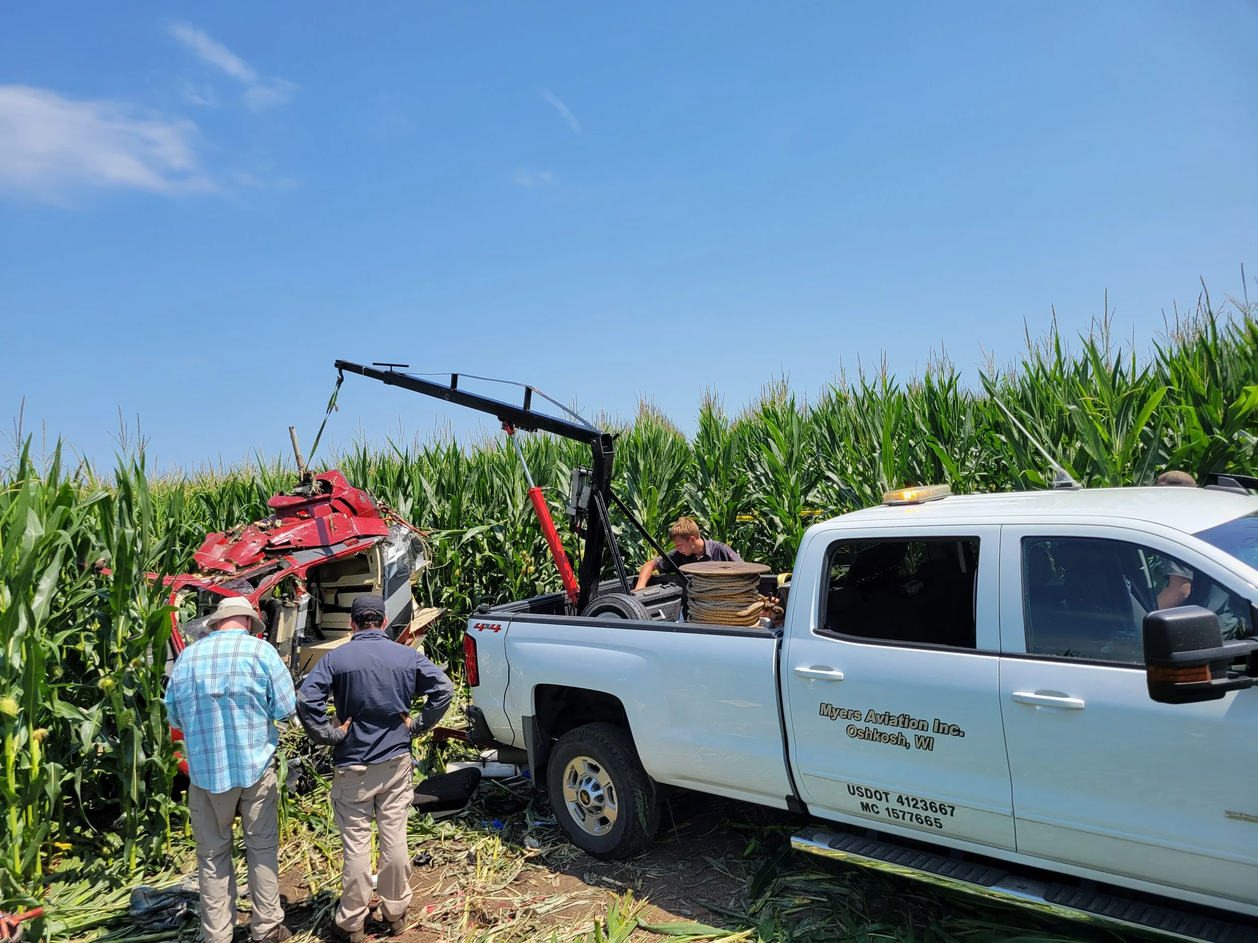 A car accident scene involving a red vehicle and a white vehicle with Myers Aviation Inc. on it, surrounded by green crops and a blue sky. Several people are inspecting the crash site with tools and equipment.