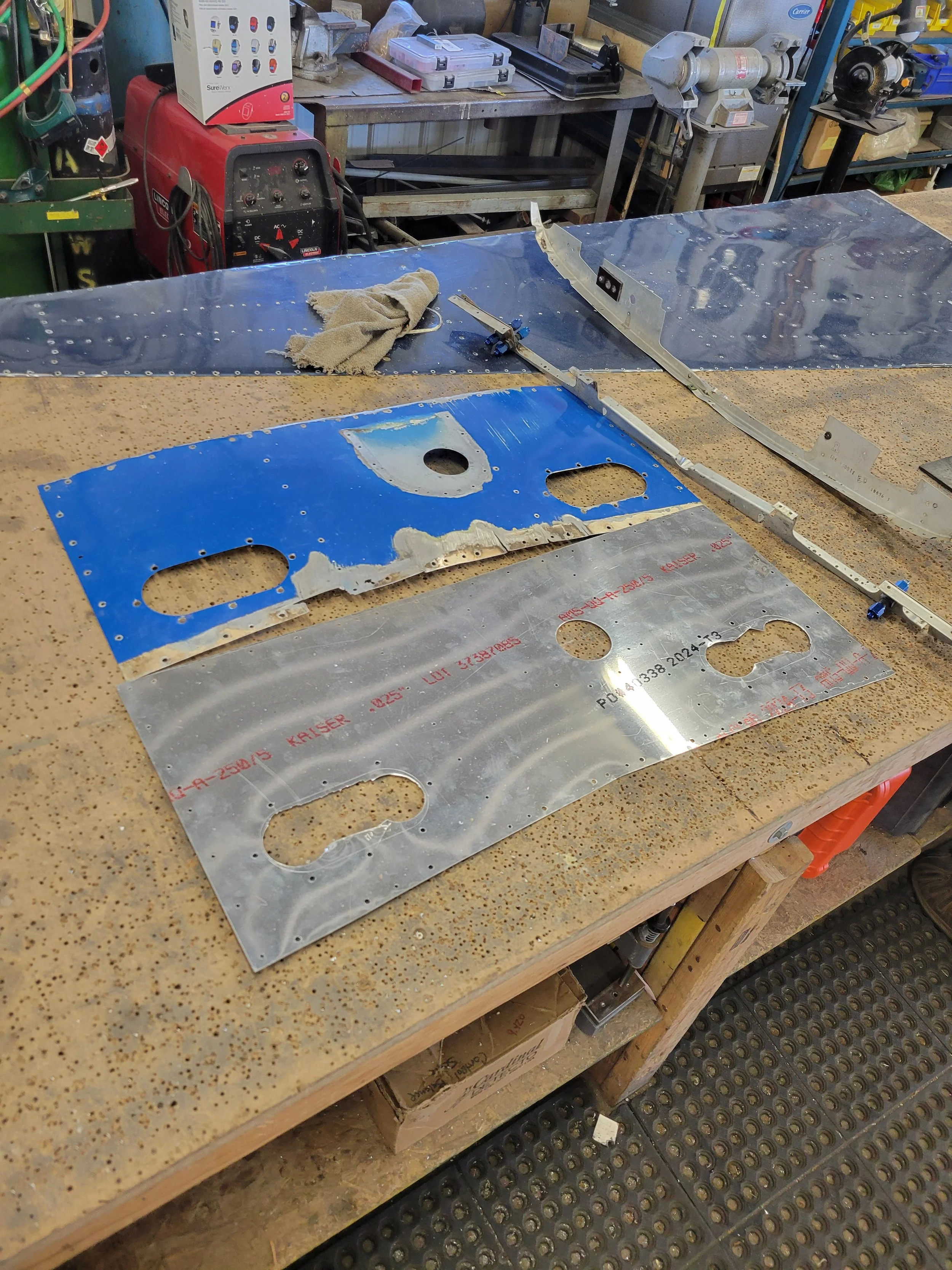 Workstation with metal pieces and tools, including a blue and silver metal panel with cutouts, a cloth, and a small metal component, surrounded by workshop equipment and supplies.