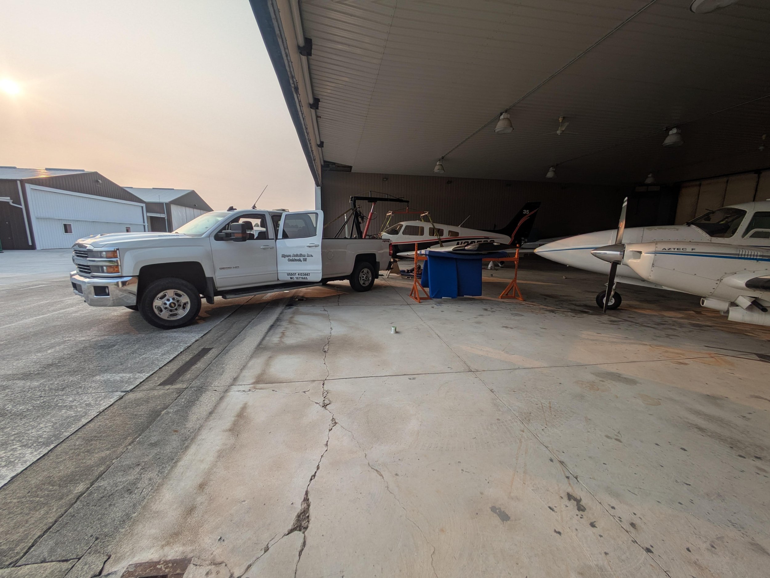 A white pickup truck parked inside an aircraft hangar with a small jet airplane in the background, and a blue trash bin in the foreground, under a corrugated metal roof with multiple lights.