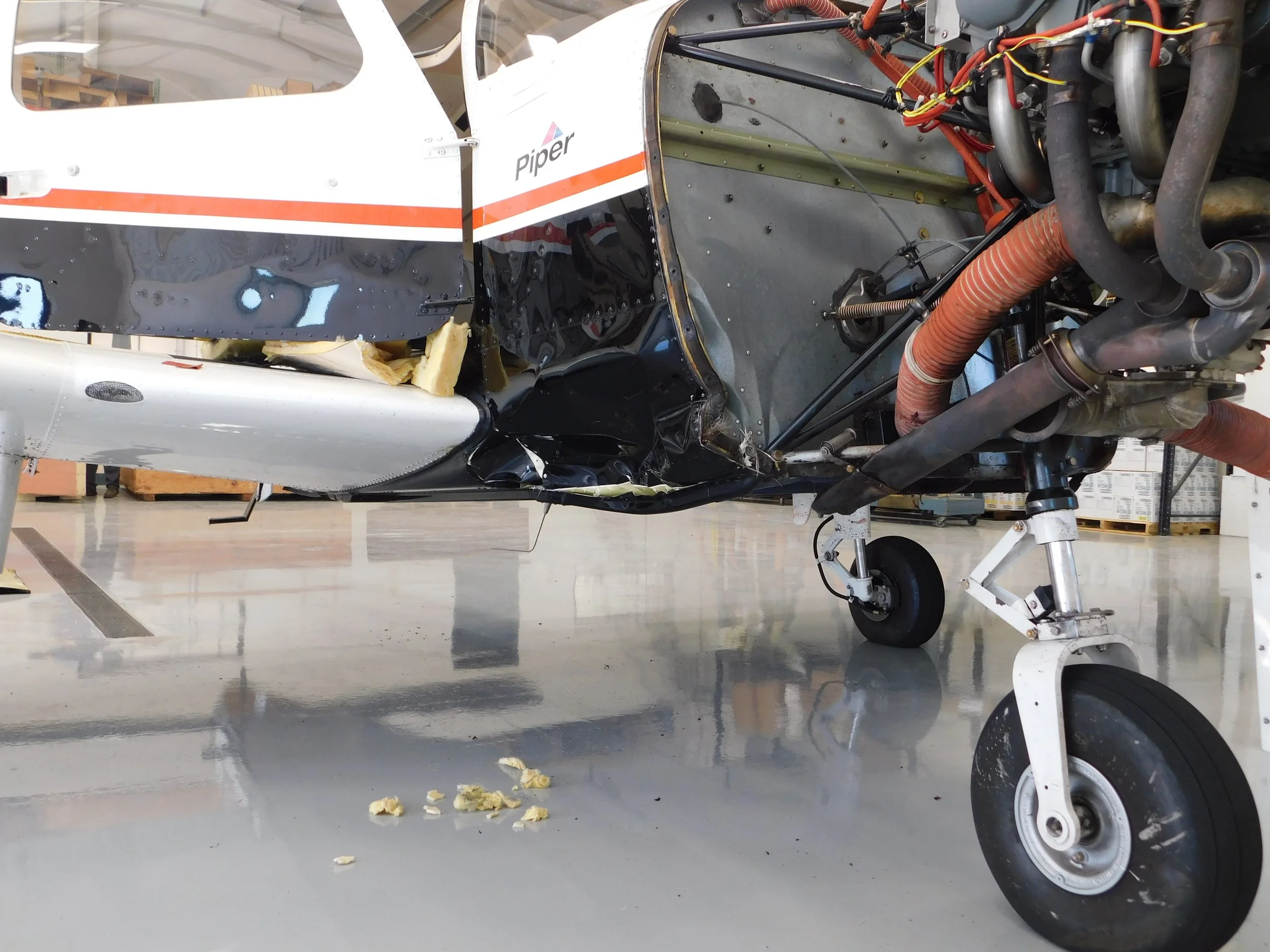 Inside view of an aircraft's cockpit showing various wires, pipes, and mechanical components, with the left side of the fuselage visible, featuring the word 'Piper' and orange and white stripes.
