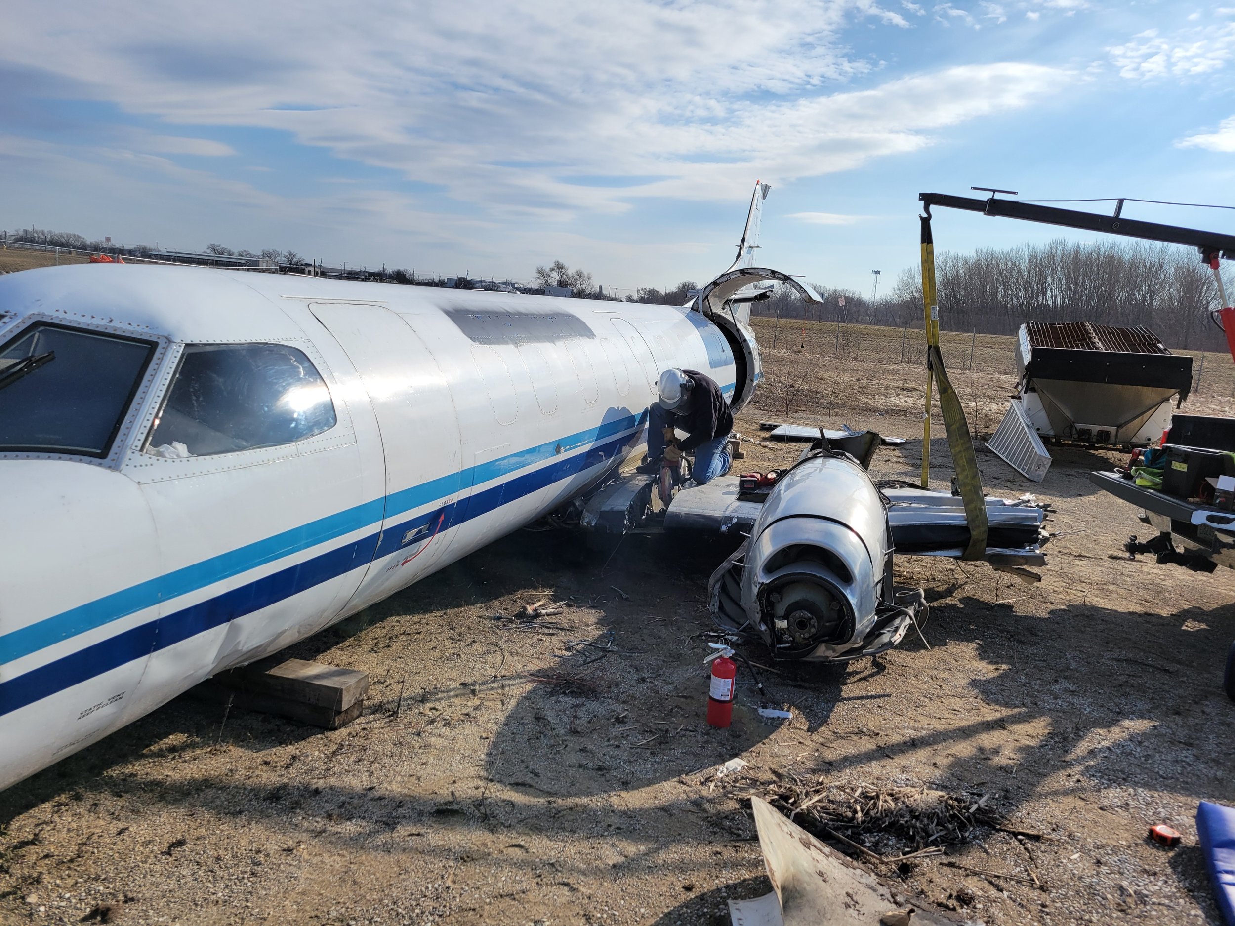 A plane fuselage with damage, being repaired outdoors with various tools and equipment around, under a cloudy sky.