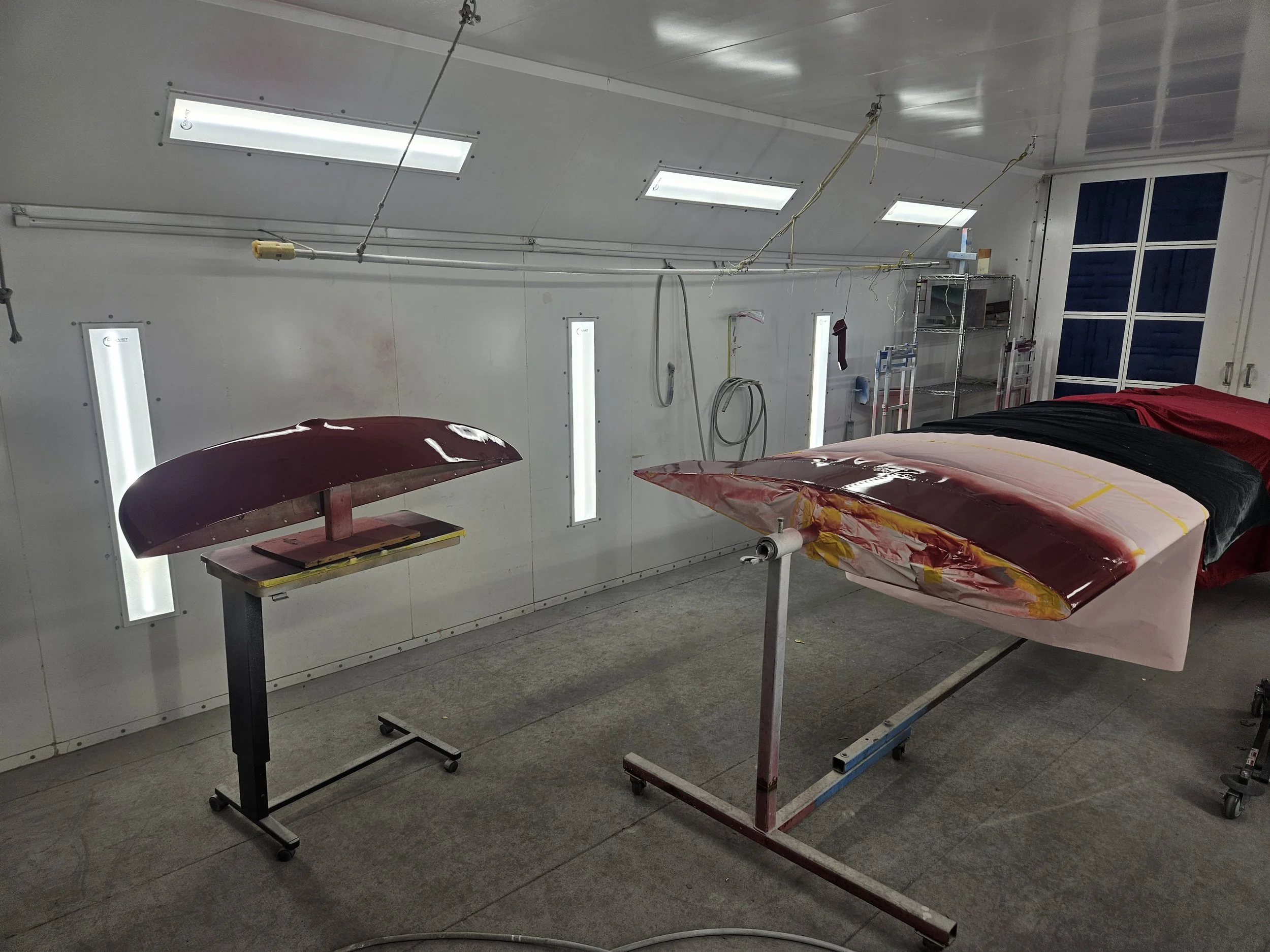 Indoor workshop with two plane skins covered in protective film, a work table, and storage shelves along the wall, with ceiling lights and tools hanging from the wall.