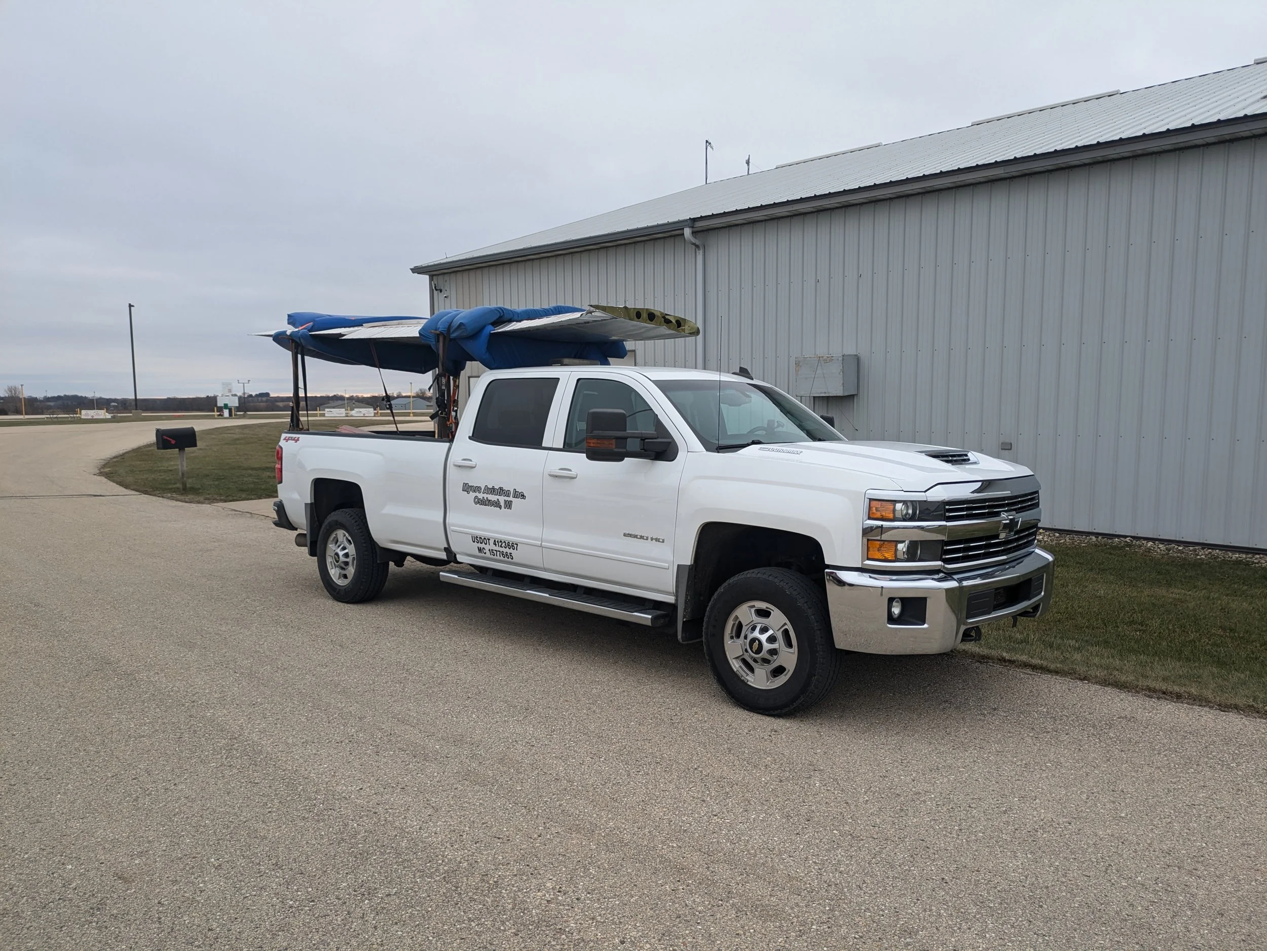 White pickup truck parked outside a large metal building, with a partly covered boat on its bed.