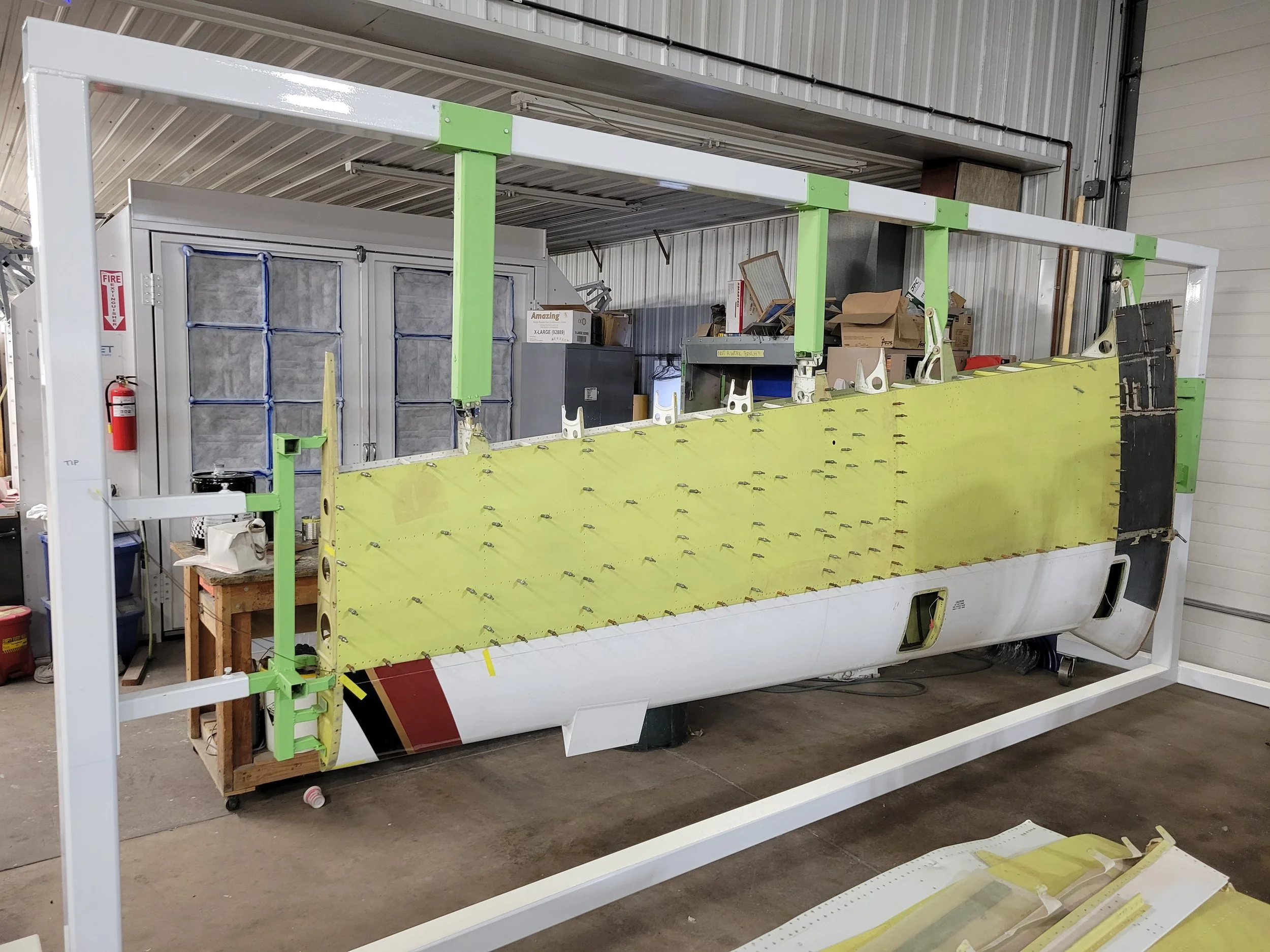 The image shows a partially assembled large model or structure, likely a boat or aircraft, inside a warehouse or workshop. The framework is white with green joints, and parts of the yellow surface are visible, with hooks or fasteners attached. The background contains storage boxes, tools, and industrial equipment.