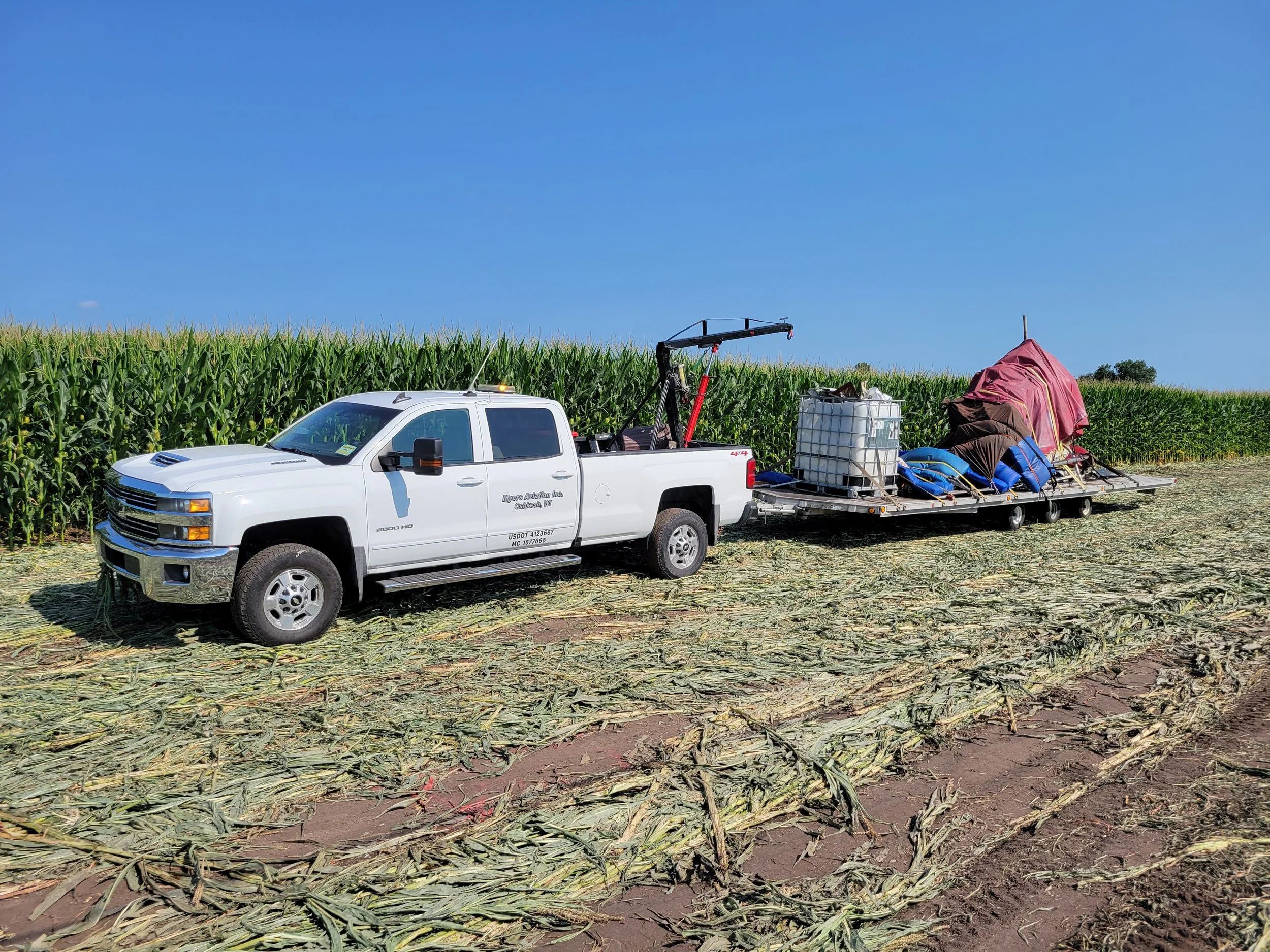A white pickup truck parked in front of a cornfield with equipment and supplies loaded on a flatbed trailer behind it.