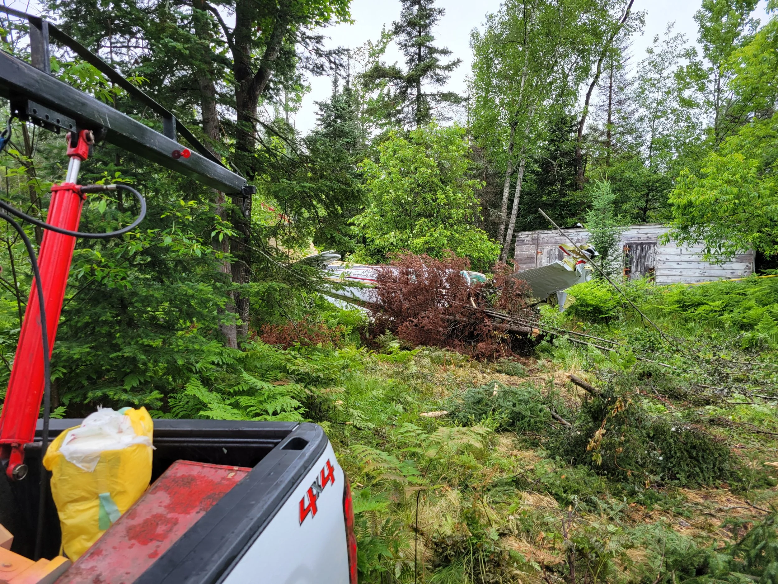 Scene of a forest with fallen trees and debris, with a damaged shed visible in the background. In the foreground, a red equipment lift with wires and a yellow bag on it.