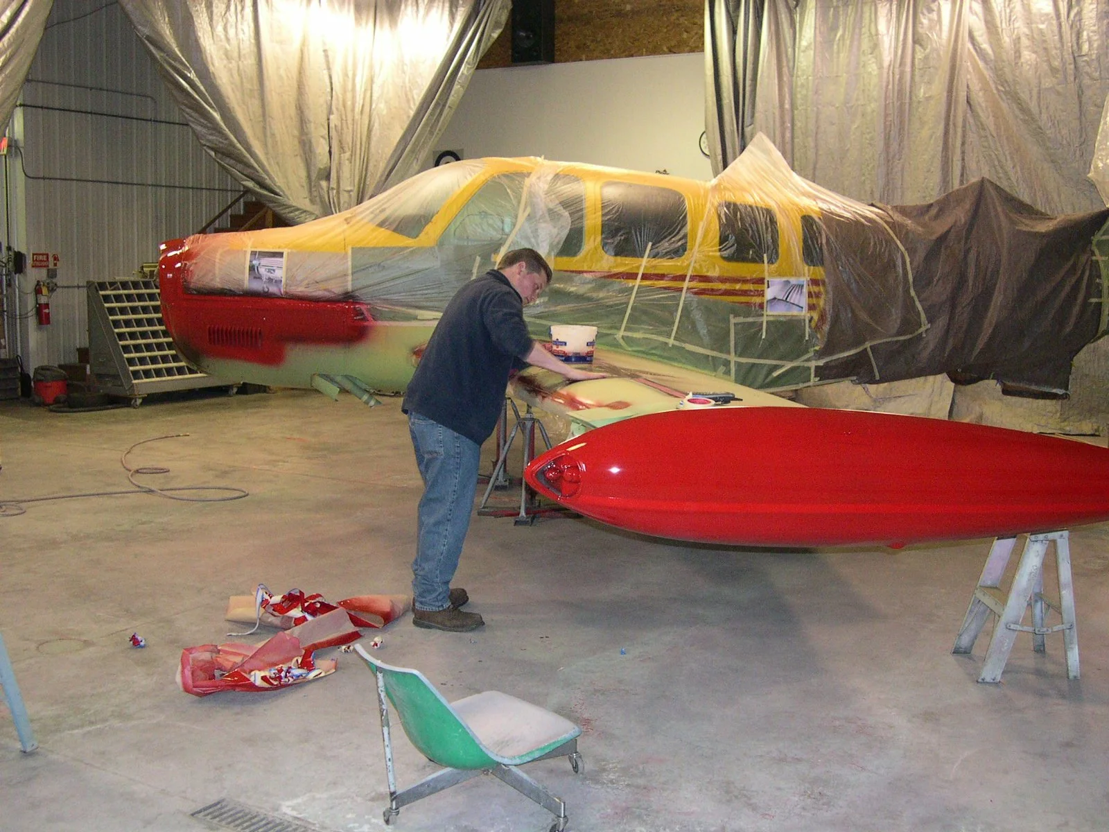 A person working on a small aircraft in a hangar, with parts of the plane covered in protective plastic and masking tape, and a red kayak on a stand nearby.